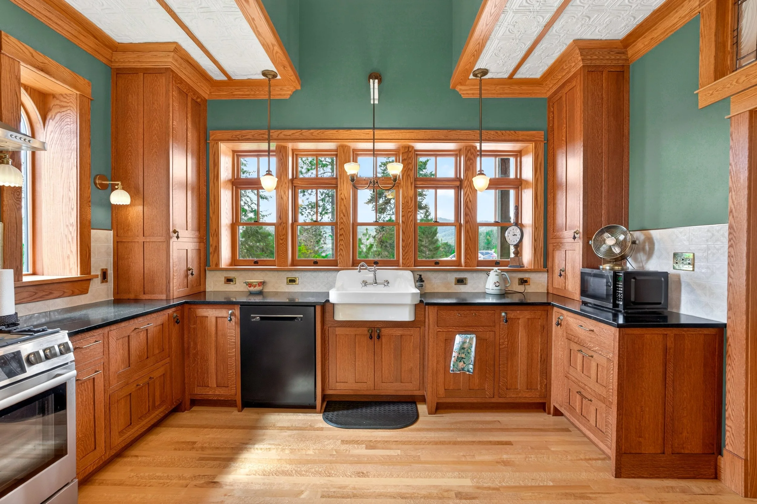 Chewelah Real Estate Photography - A kitchen with wooden cabinets, a white farmhouse sink under a large window, granite countertops, a stainless steel stove, and small appliances including a microwave and toaster on the counter.