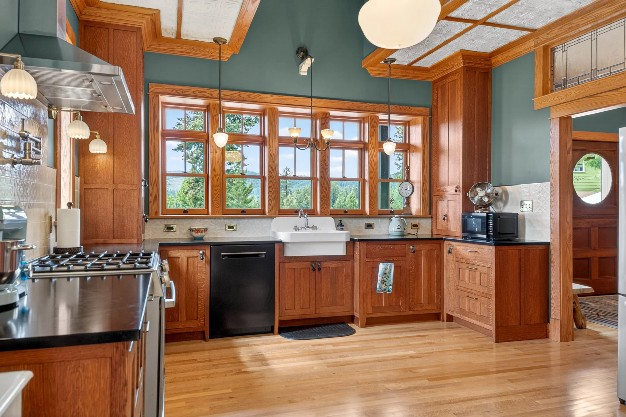Chewelah Real Estate Photography - A kitchen with wooden cabinets and large windows with a scenic view outside. It has a white farmhouse sink, black countertops, and various appliances including a microwave and a fan. 
