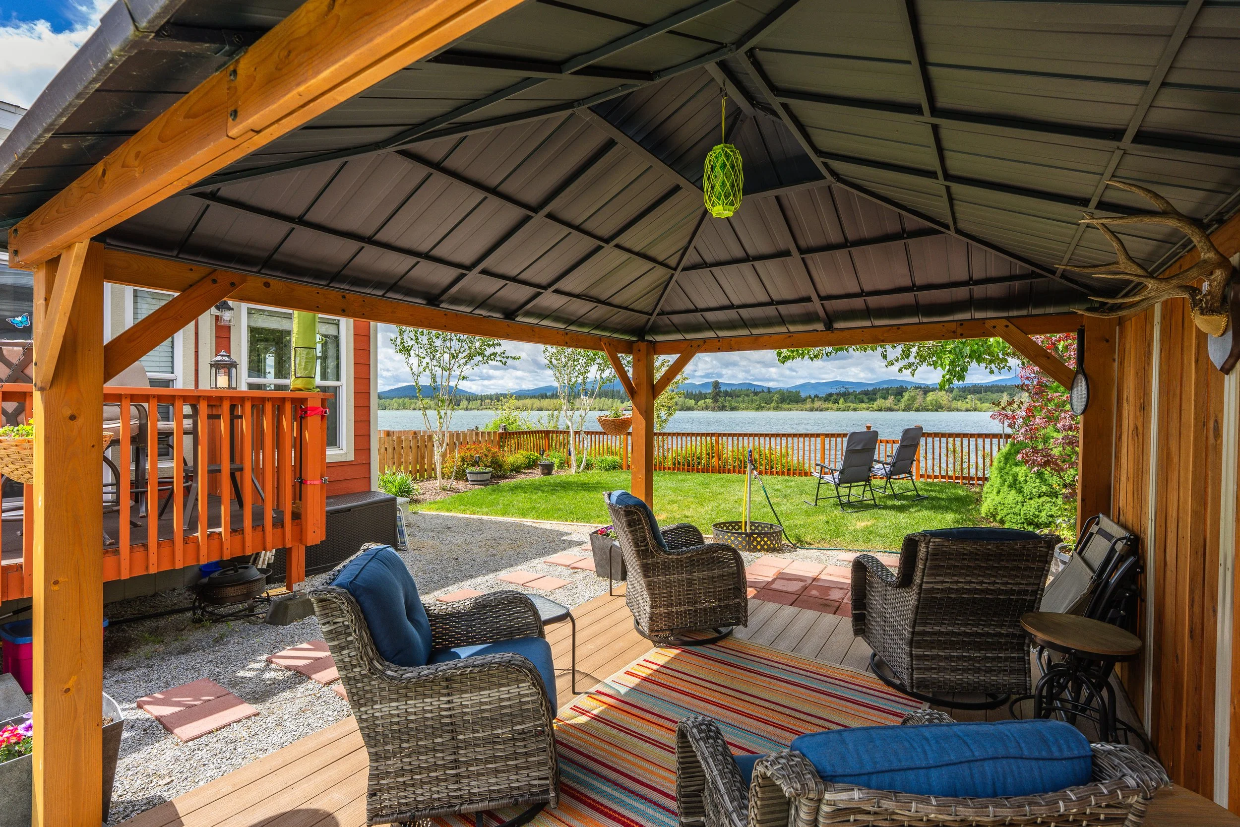 Pend Oreille County Real Estate Photography - Covered outdoor patio with wicker chairs, a colorful striped rug, a wooden deck, a view of a lake and trees in the background, and a hunting antler wall decoration.