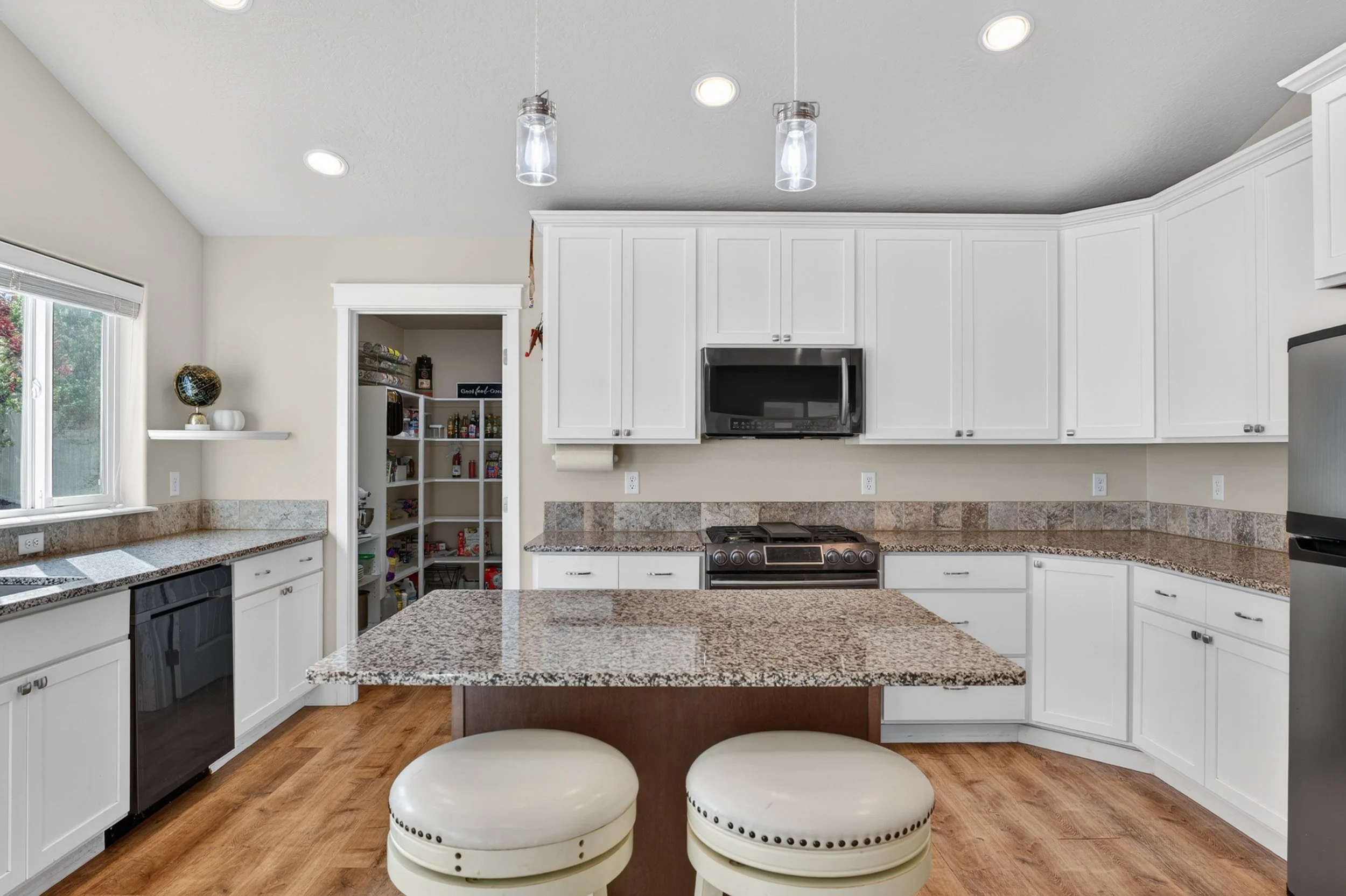 Spokane Real Estate Photography - A modern kitchen with white cabinets, granite countertops, a stainless steel microwave, and a stove, with a kitchen island and two cream-colored stools.