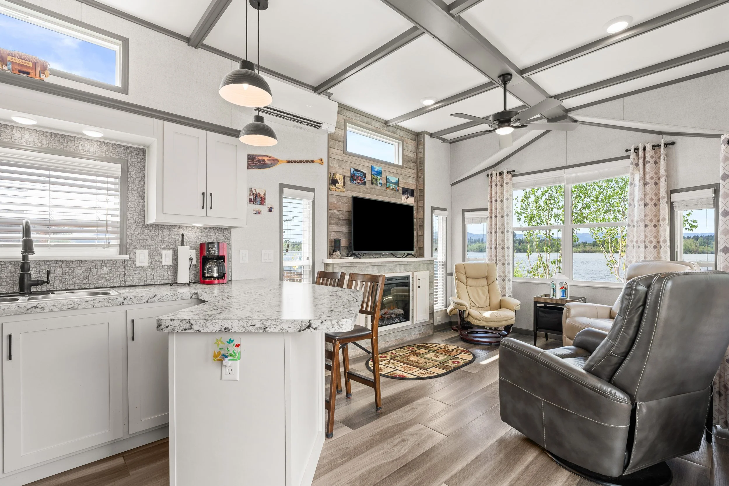 Pend Oreille County Real Estate Photography - Open-concept living room and kitchen area with large windows, gray walls, white cabinetry, a granite kitchen island, and modern furniture including beige and black chairs.