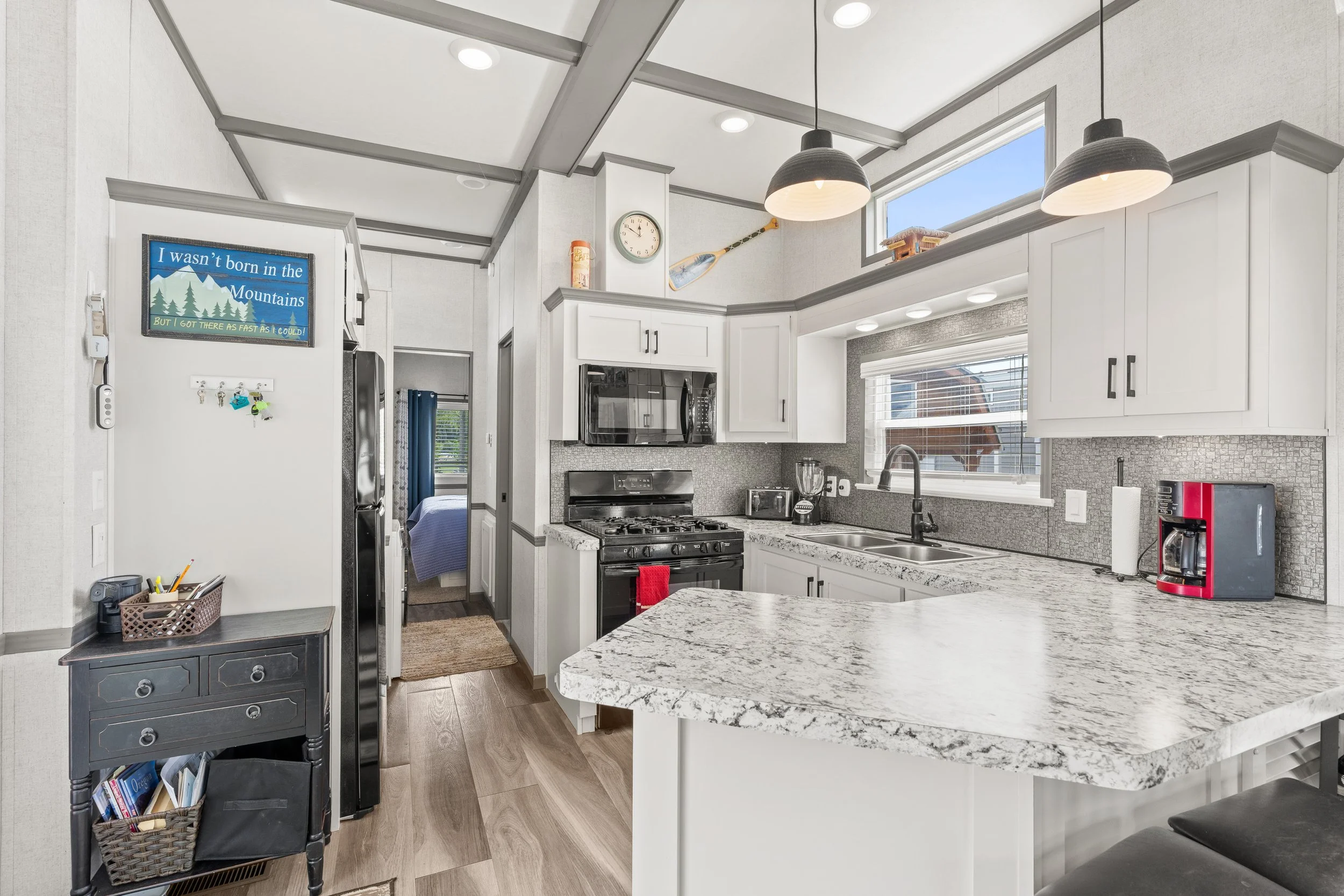 Pend Oreille County Real Estate Photography - Bright kitchen with white cabinets, marble countertops, stainless steel appliances, and a window above the sink. Decor includes a wall clock, painting, and two pendant lights.