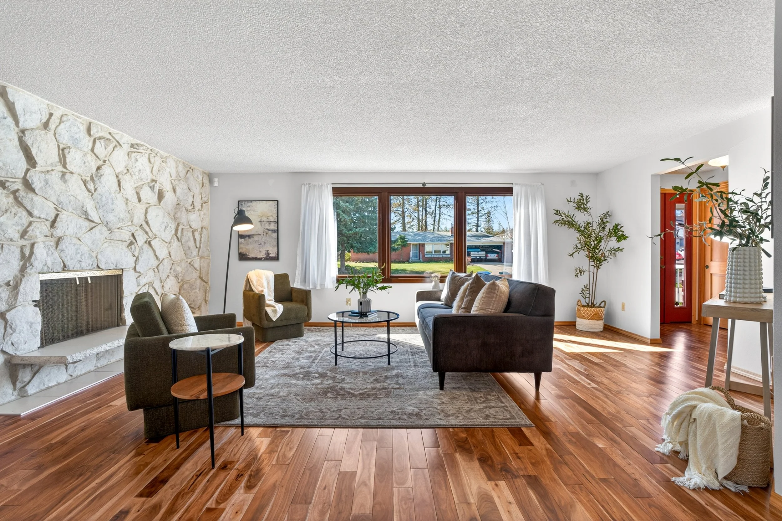 Spokane Real Estate Photography - Living room with stone fireplace, large window with white curtains, and hardwood floors. Contains sofa, armchairs, side table, plants, and artwork.