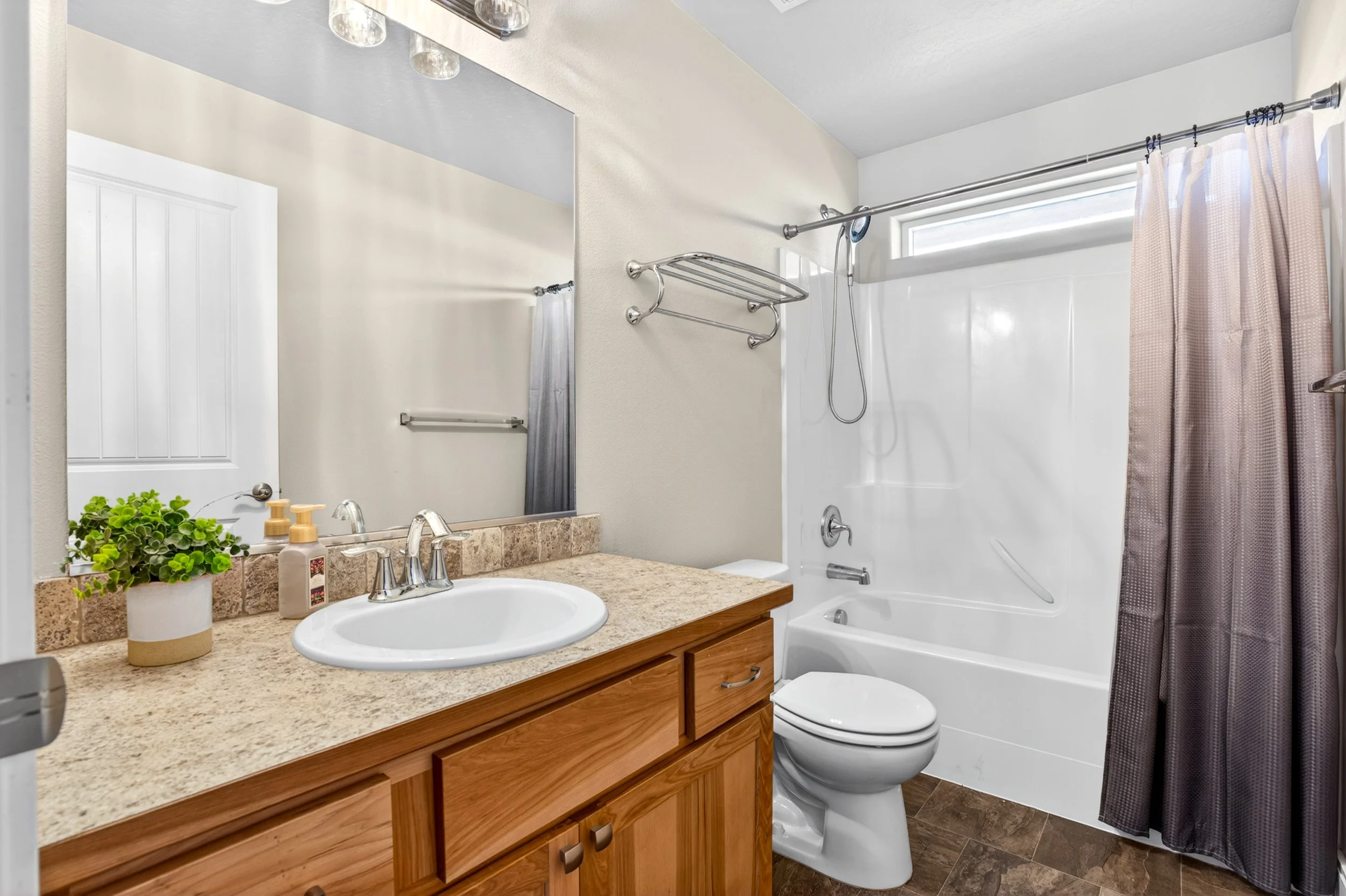 Spokane Real Estate Photography - A bathroom with a vanity sink, mirror, and countertop with a plant and soap dispensers, a toilet, a bathtub with a showerhead, a window with a curtain, and wall-mounted shelves.