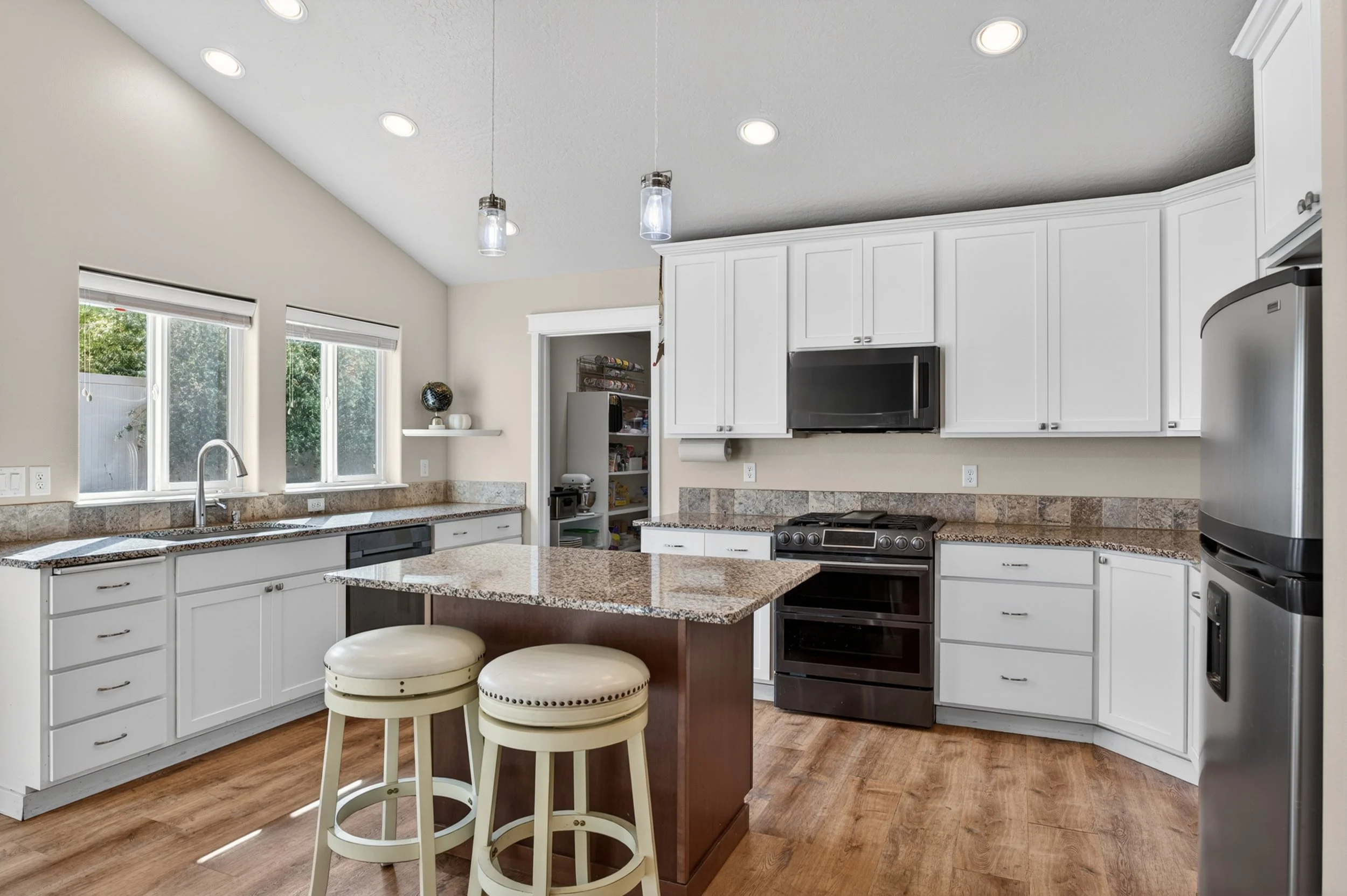 Spokane Real Estate Photography - A modern kitchen with white cabinets, granite countertops, stainless steel appliances, and a small island with two cream-colored barstools. Large windows with white blinds let in natural light.