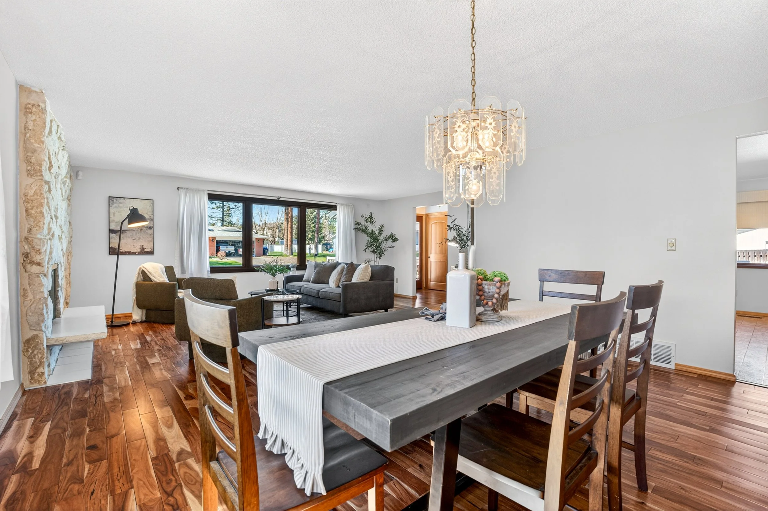 Spokane Real Estate Photography - Open living and dining room with hardwood floors, a sectional sofa, a large window, a dining table with chairs, and a decorative chandelier.