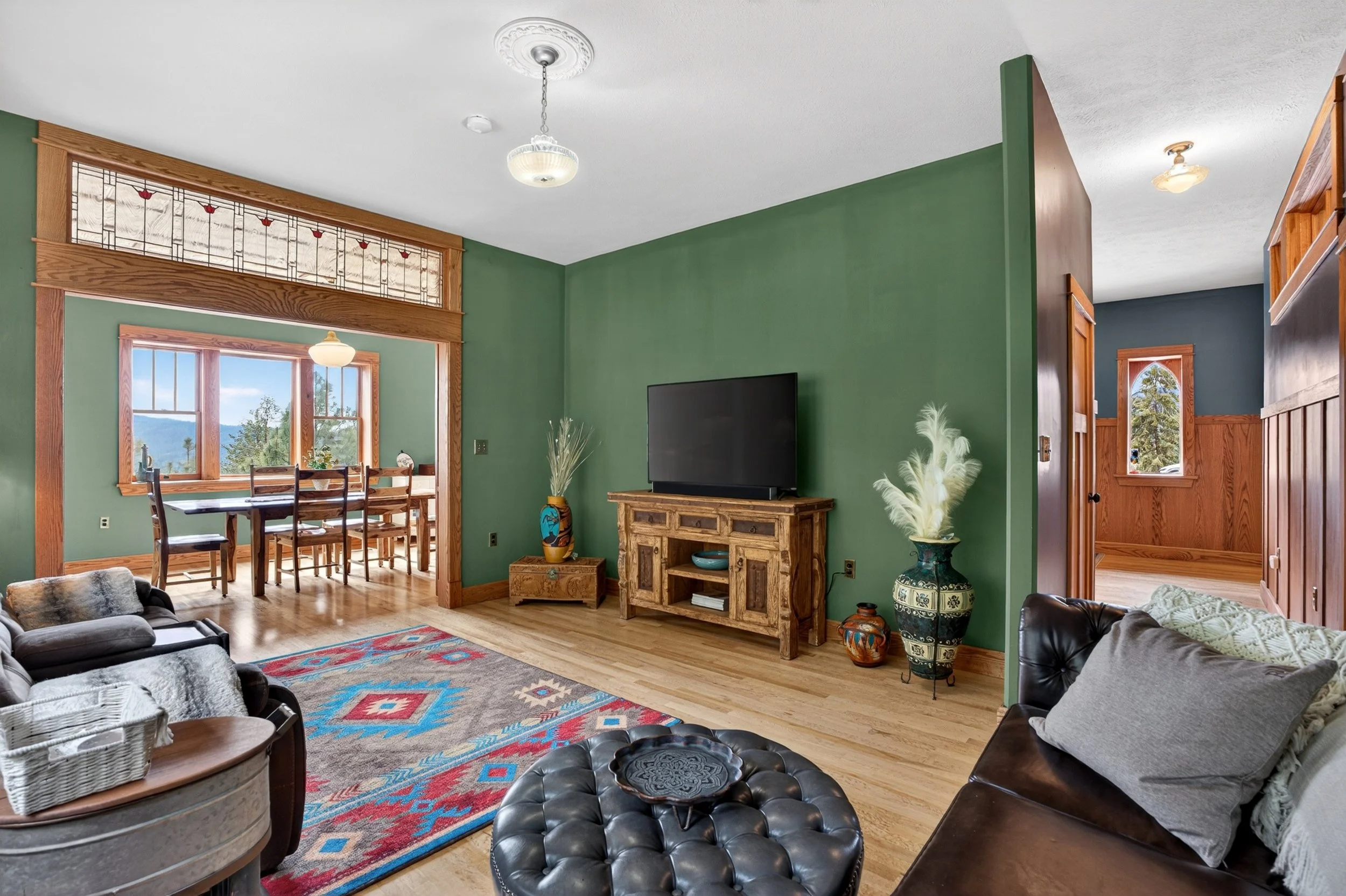 Chewelah Real Estate Photography - A living room with wooden floors, green walls, and various furniture including couches, a wooden TV stand, a patterned area rug, and decorative vases.