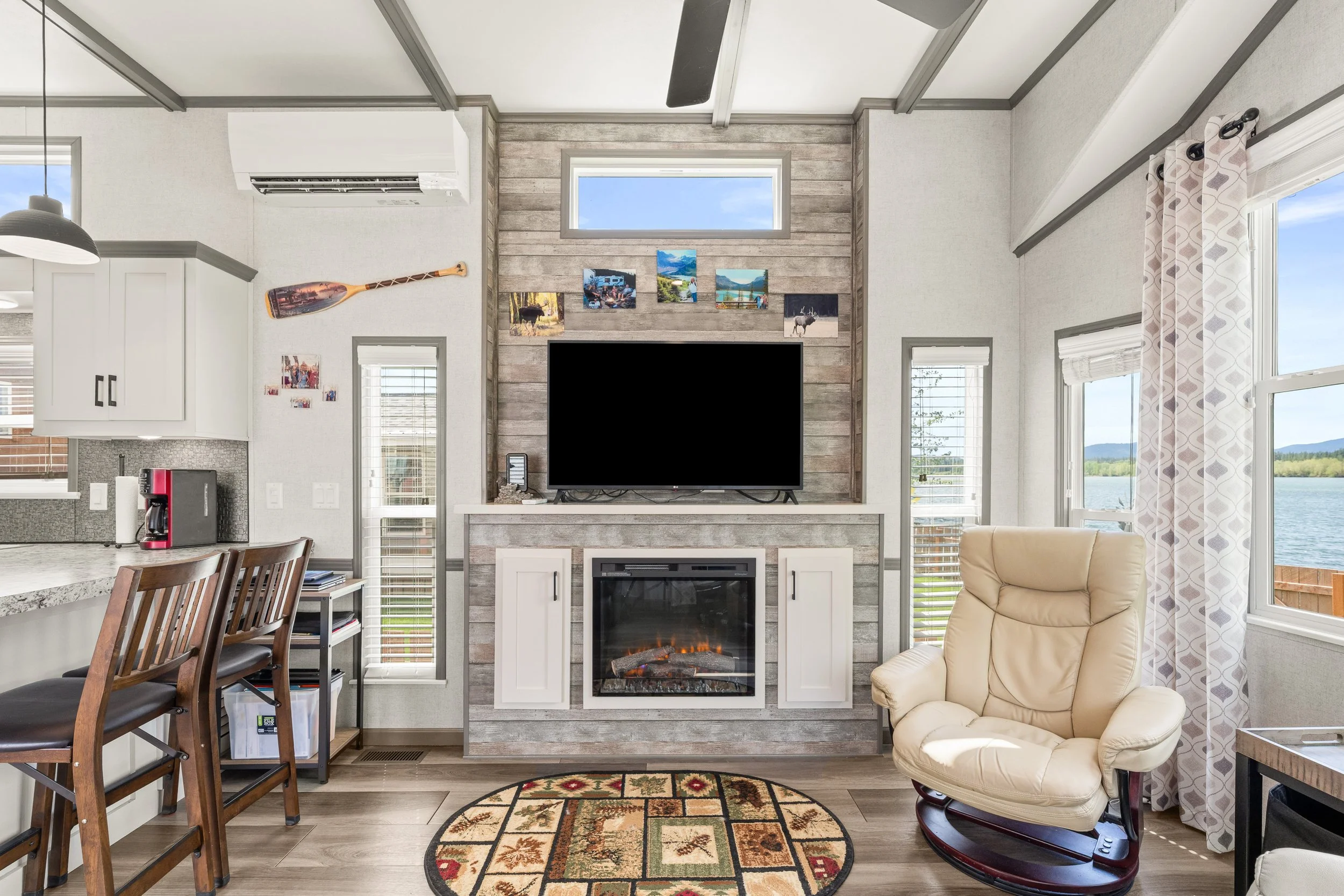 Pend Oreille County Real Estate Photography - Living room with a fireplace, television, large windows, a beige recliner chair, and a patterned rug on wooden flooring.