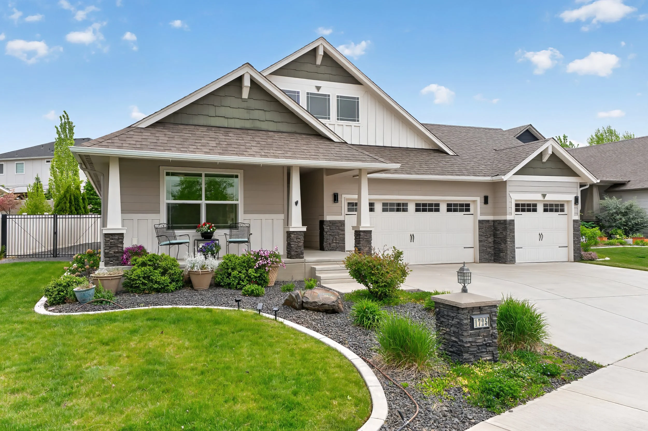 Spokane Real Estate Photography - A suburban house with a well-maintained front yard, concrete driveway, and two-car garage. The house has a grey exterior with a gabled roof and a front porch with outdoor seating and potted plants. 