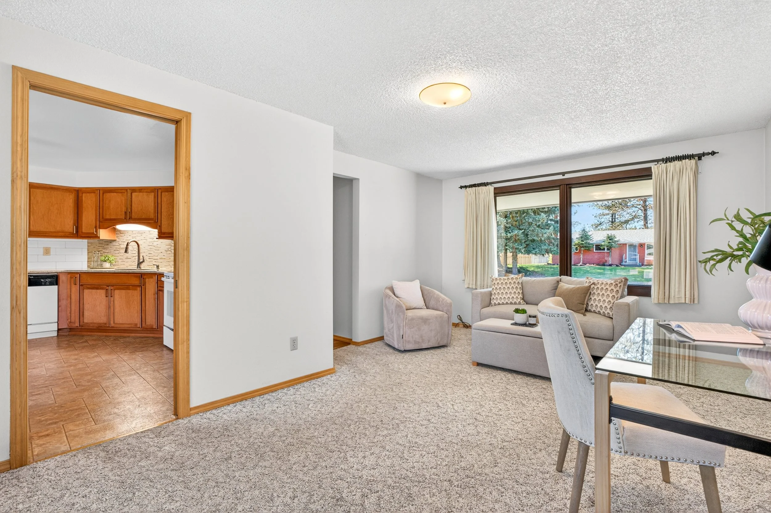 Spokane Real Estate Photography - Living room with carpeted floor, large window with curtains, beige sofa and armchair, and a view of a backyard with trees and a red house.