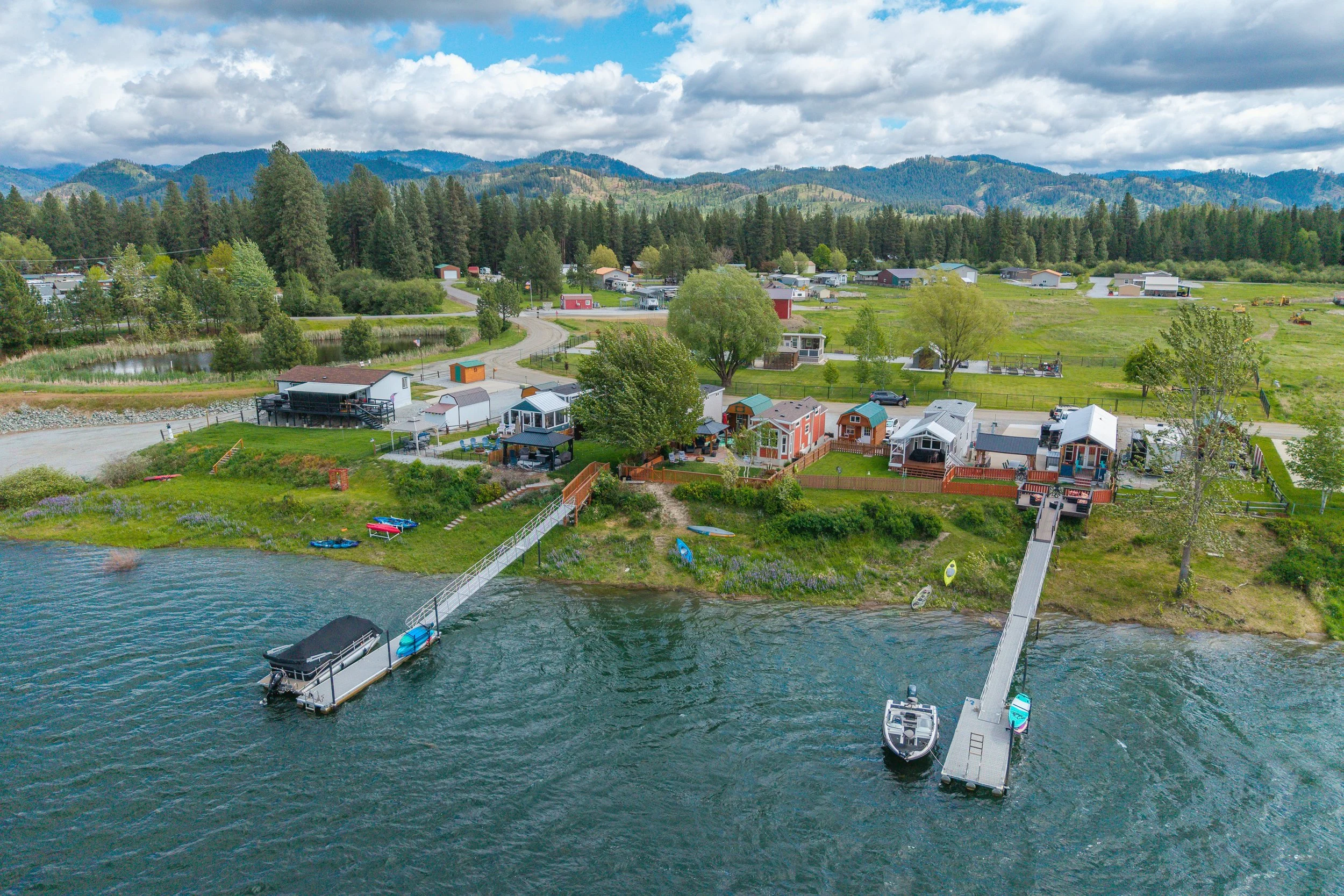 Pend Oreille County Real Estate Photography - A scenic aerial view of a lakeside community with colorful small houses near water, surrounded by green trees and mountains under a partly cloudy sky.