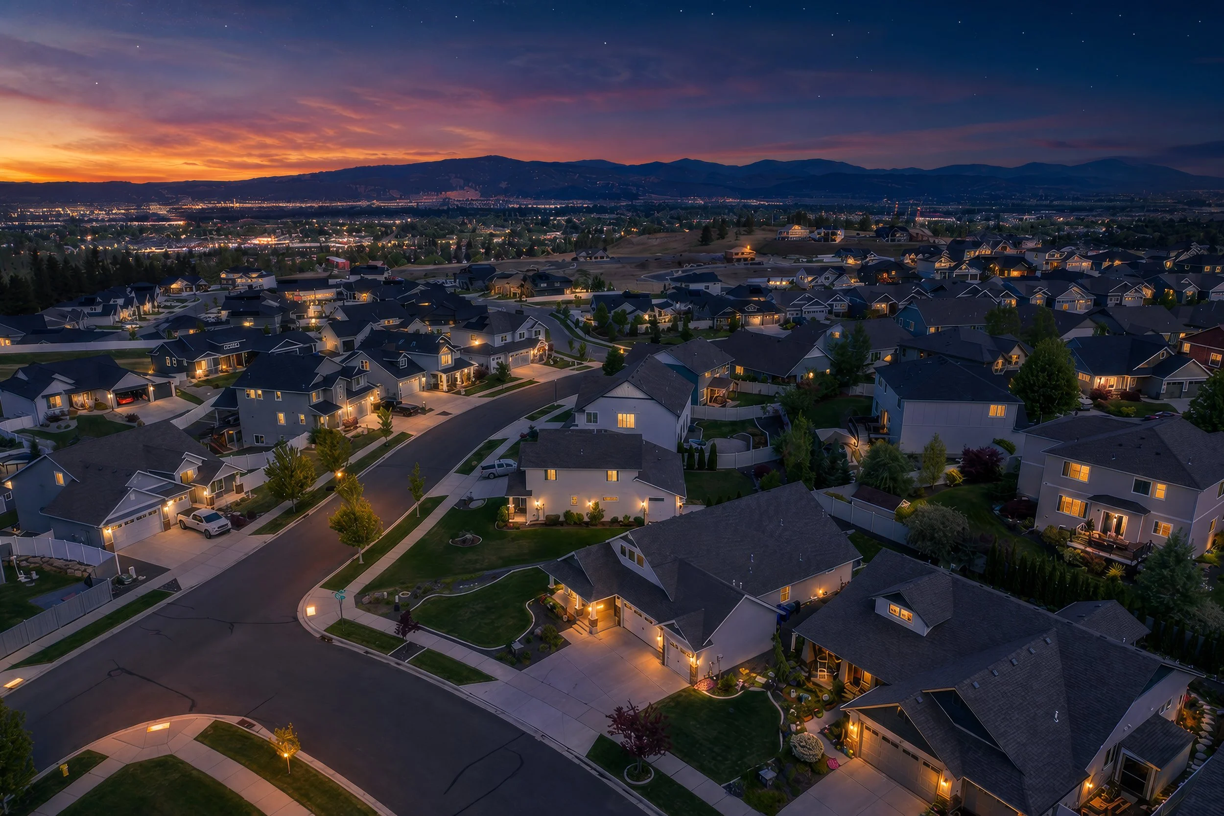 Spokane Valley Real Estate Photography - Virtual twilight of aerial view of a suburban neighborhood at dusk with houses, streets, and trees illuminated by outdoor lights, and a mountain range in the background under a colorful sunset sky.