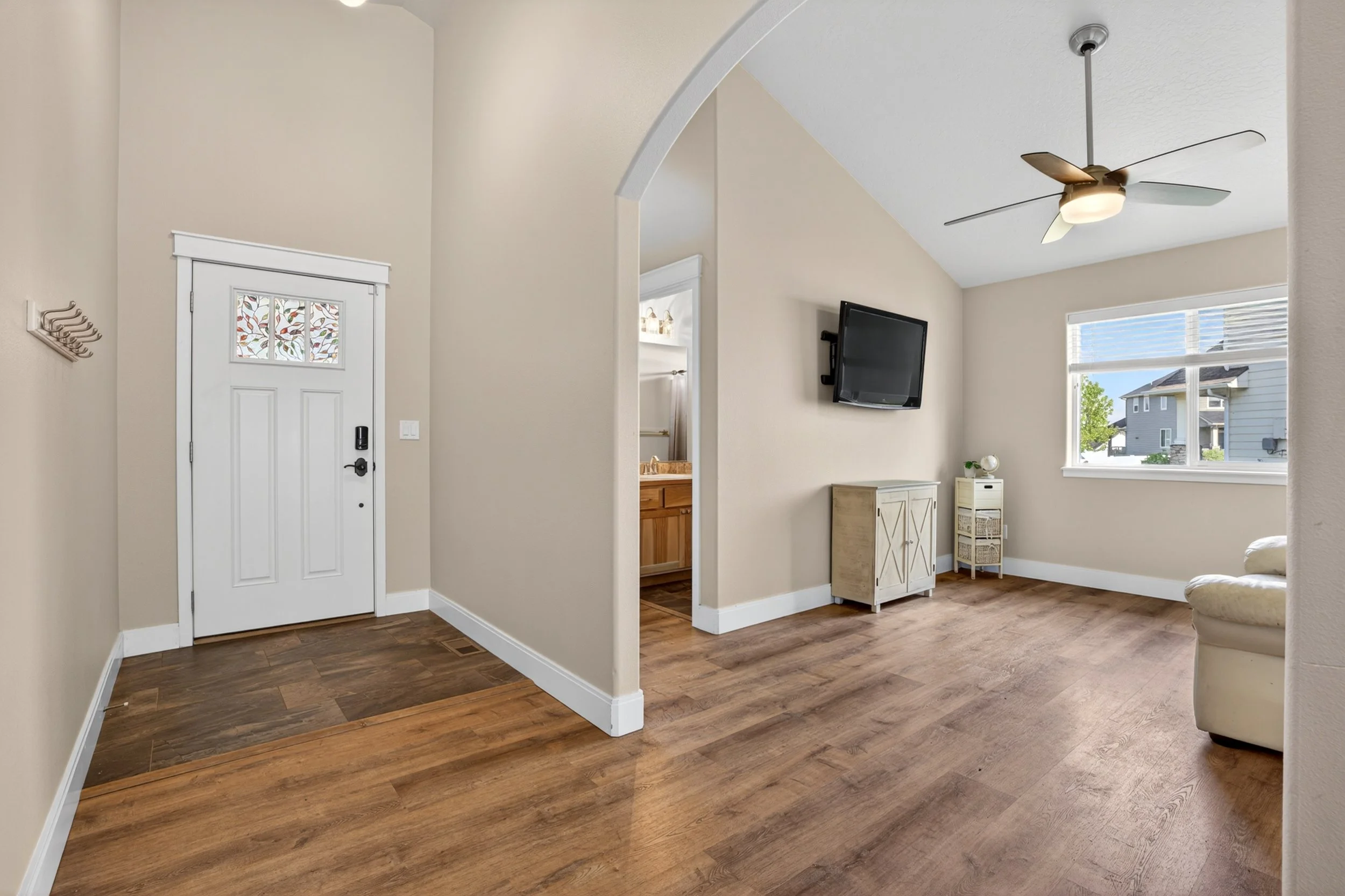 Spokane Real Estate Photography - Living room with beige walls, hardwood flooring, a ceiling fan, a wall-mounted TV, a window with a view of neighboring houses, and a small cabinet under the TV.
