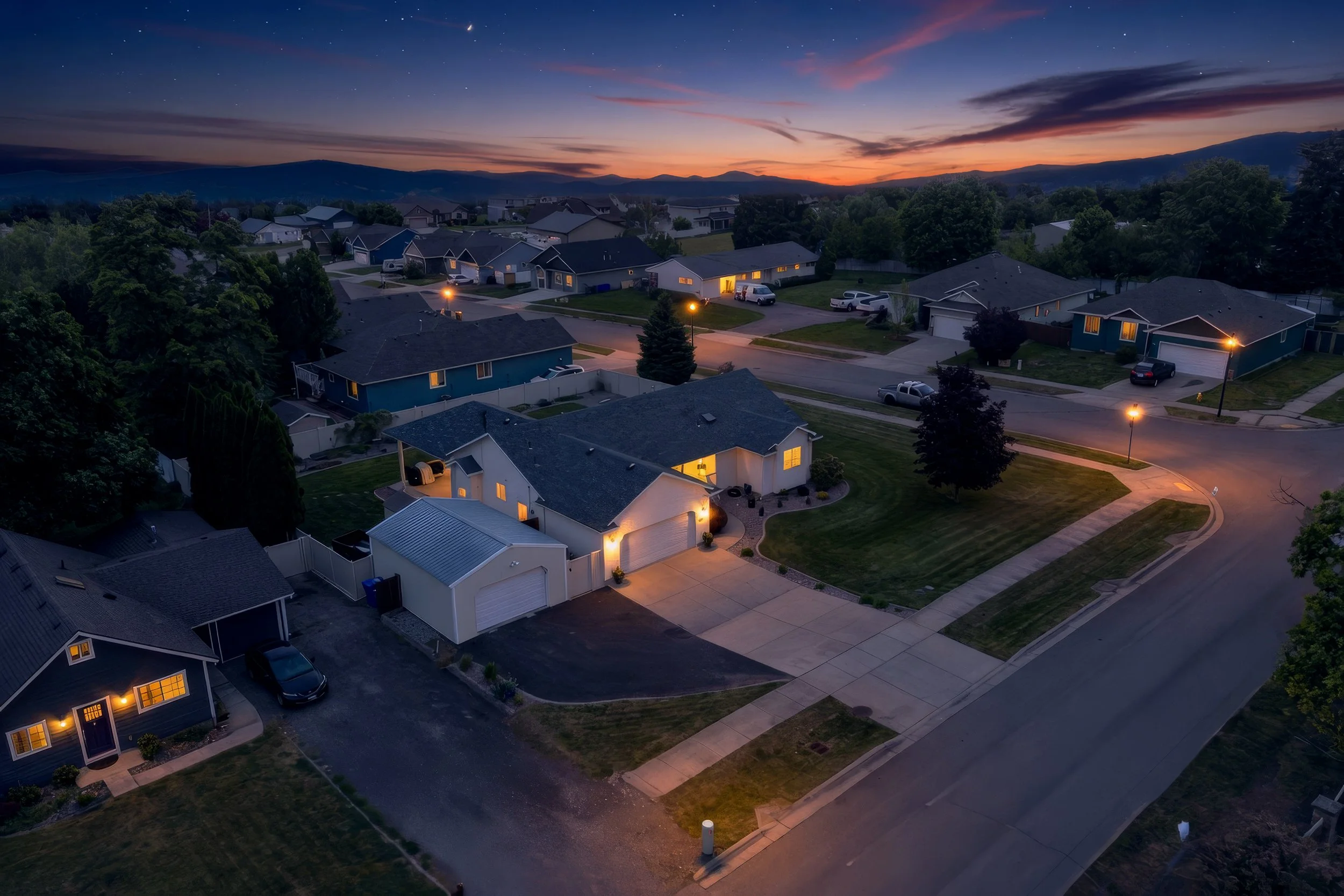 Spokane Real Estate Photography - Virtual twilight of a suburban neighborhood at dusk with houses, cars parked on driveways and streets, streetlights illuminating the area, and mountains in the background under a colorful sky with clouds and stars.