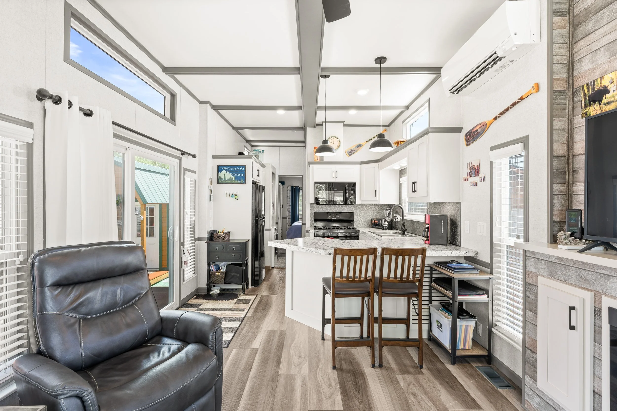 Pend Oreille County Real Estate Photography - Interior view of a modern kitchen and living area with white cabinets, stainless steel appliances, a granite countertop, and multiple windows.