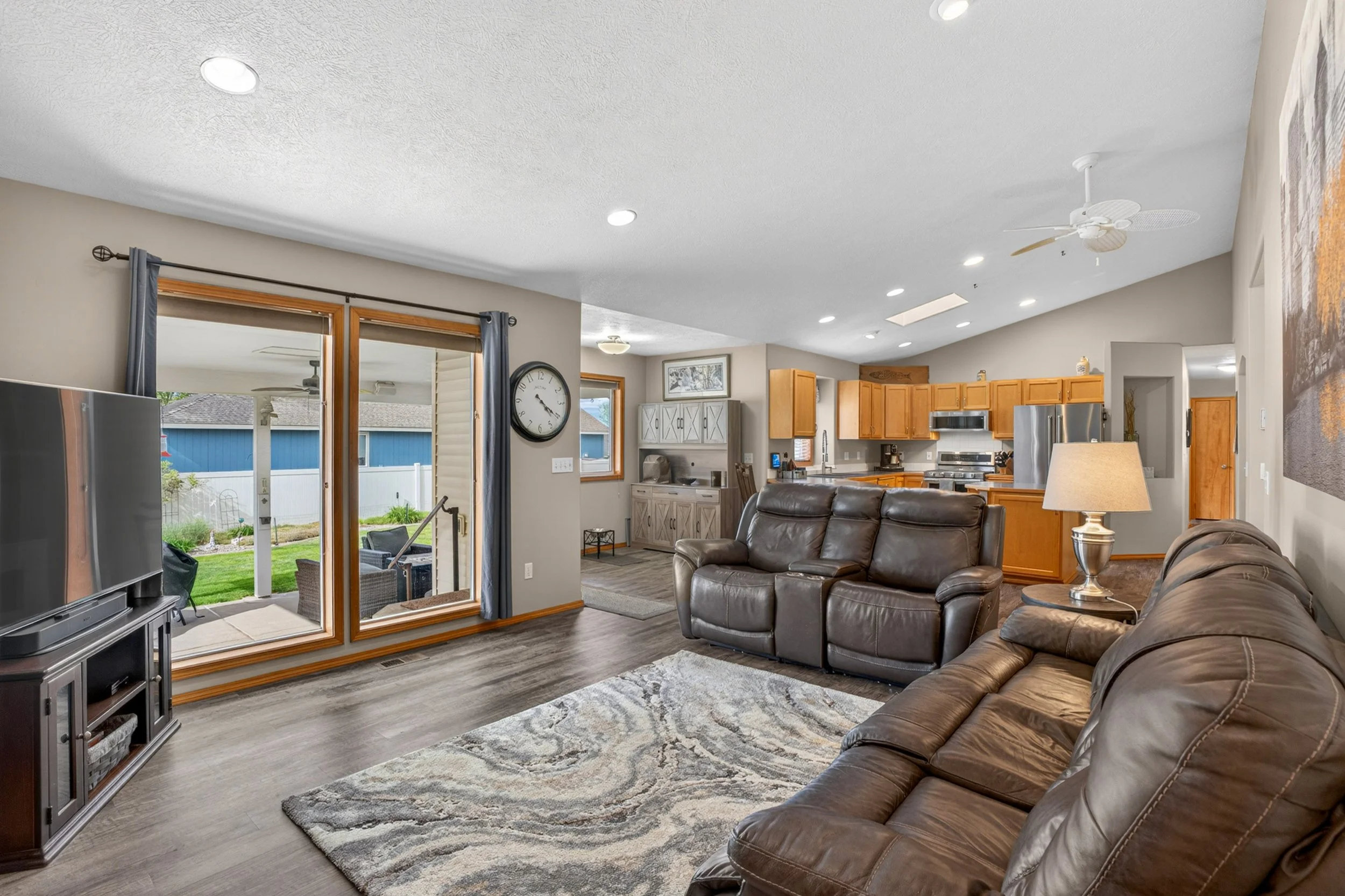 Spokane Real Estate Photography - Living room with leather sofas, a wooden cabinet, a large wall clock, sliding glass door, ceiling fan, and view of a backyard