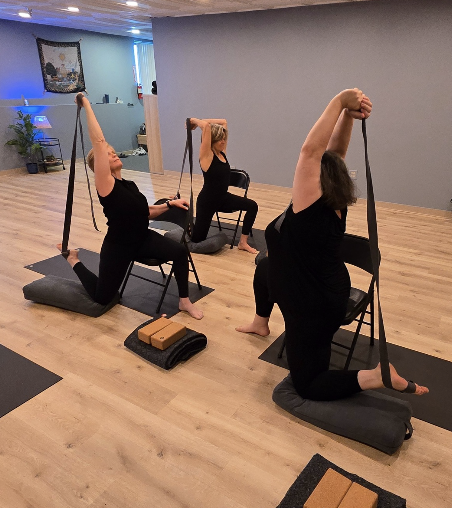 Three women practicing yoga with chairs and straps on a wooden floor in a studio.