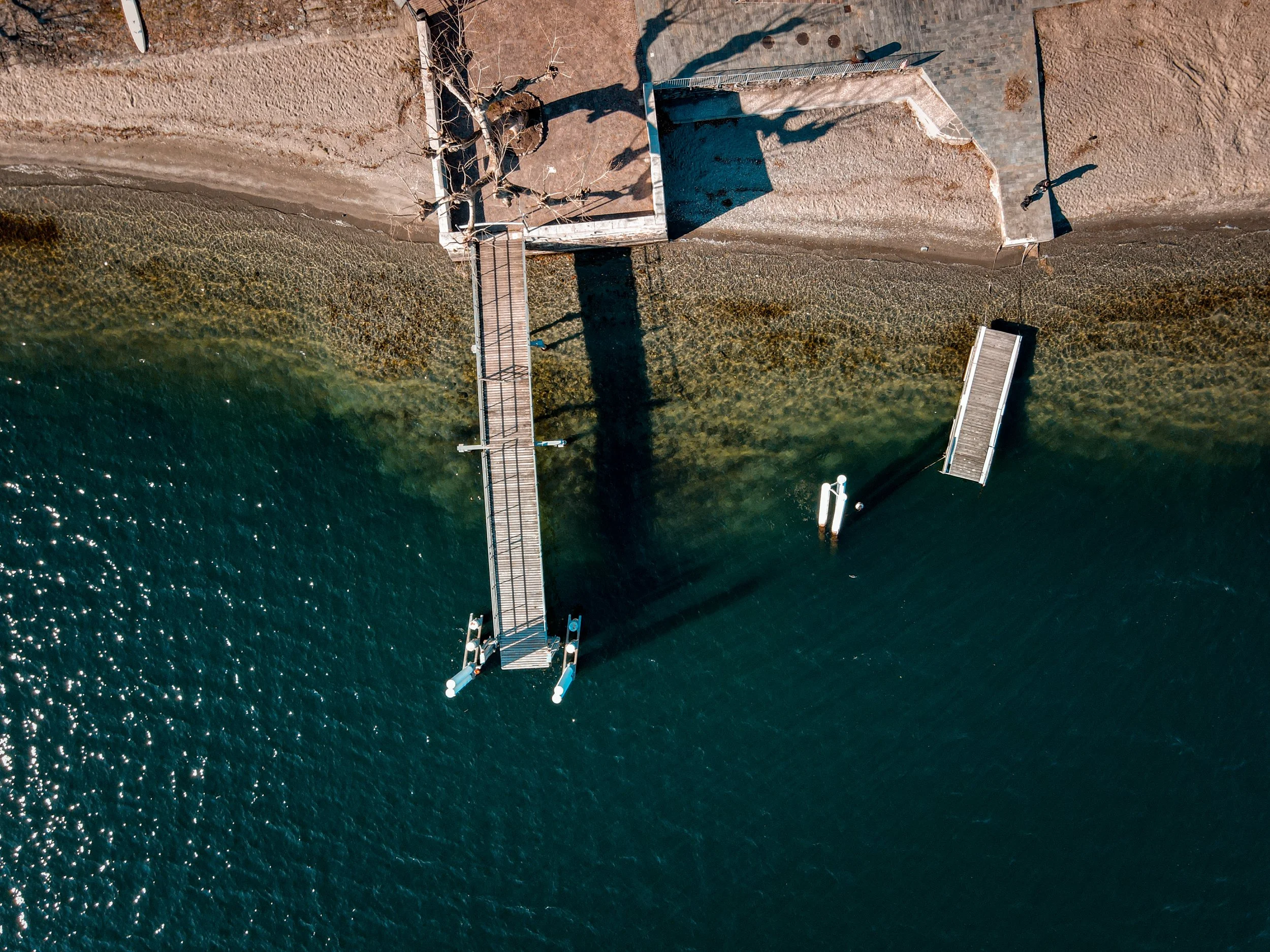 An aerial view of a waterfront with a concrete pier extending into the water, a small dock, and a sandy shoreline with some trees casting shadows.
