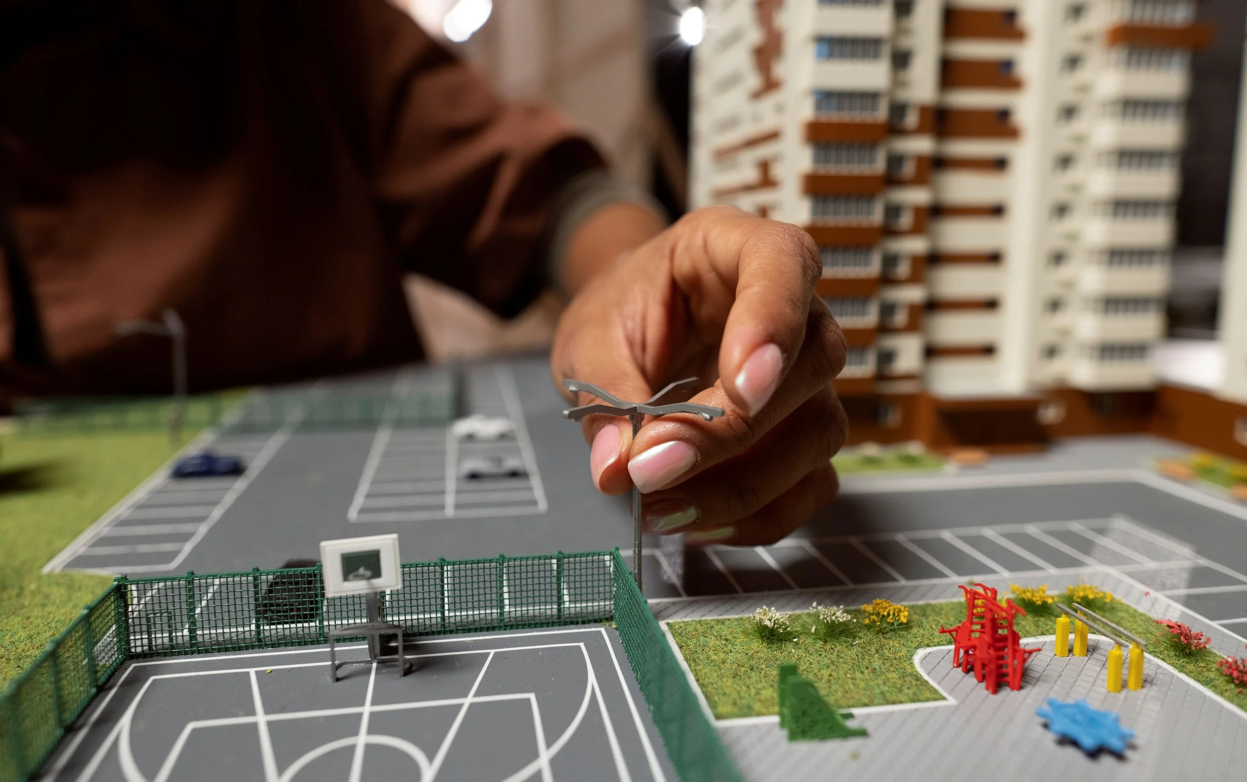 Close-up of a person's hand placing a miniature figure on a model of a sports complex, with buildings and parking lot in the background.