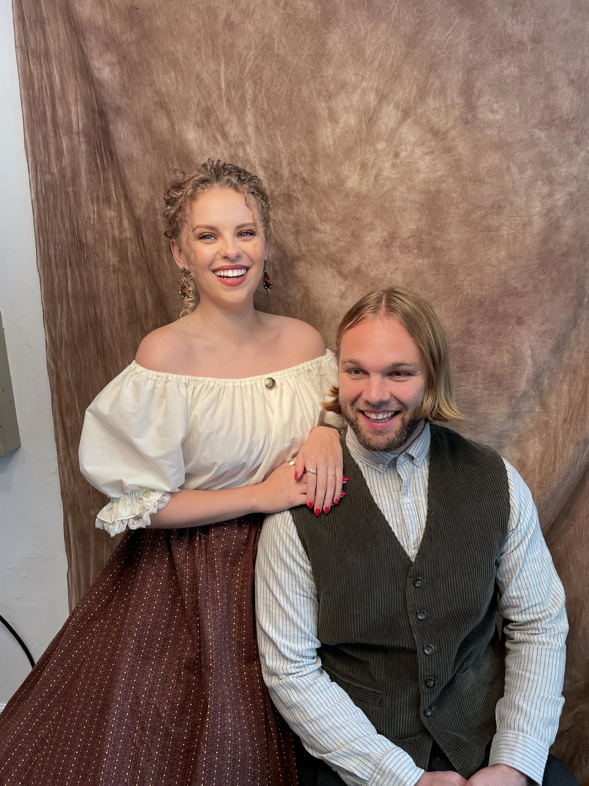 A smiling woman with curly hair, wearing earrings and a white off-shoulder top, standing behind a man with shoulder-length blond hair and a beard, who is wearing a striped shirt and dark vest, in front of a brown textured backdrop.
