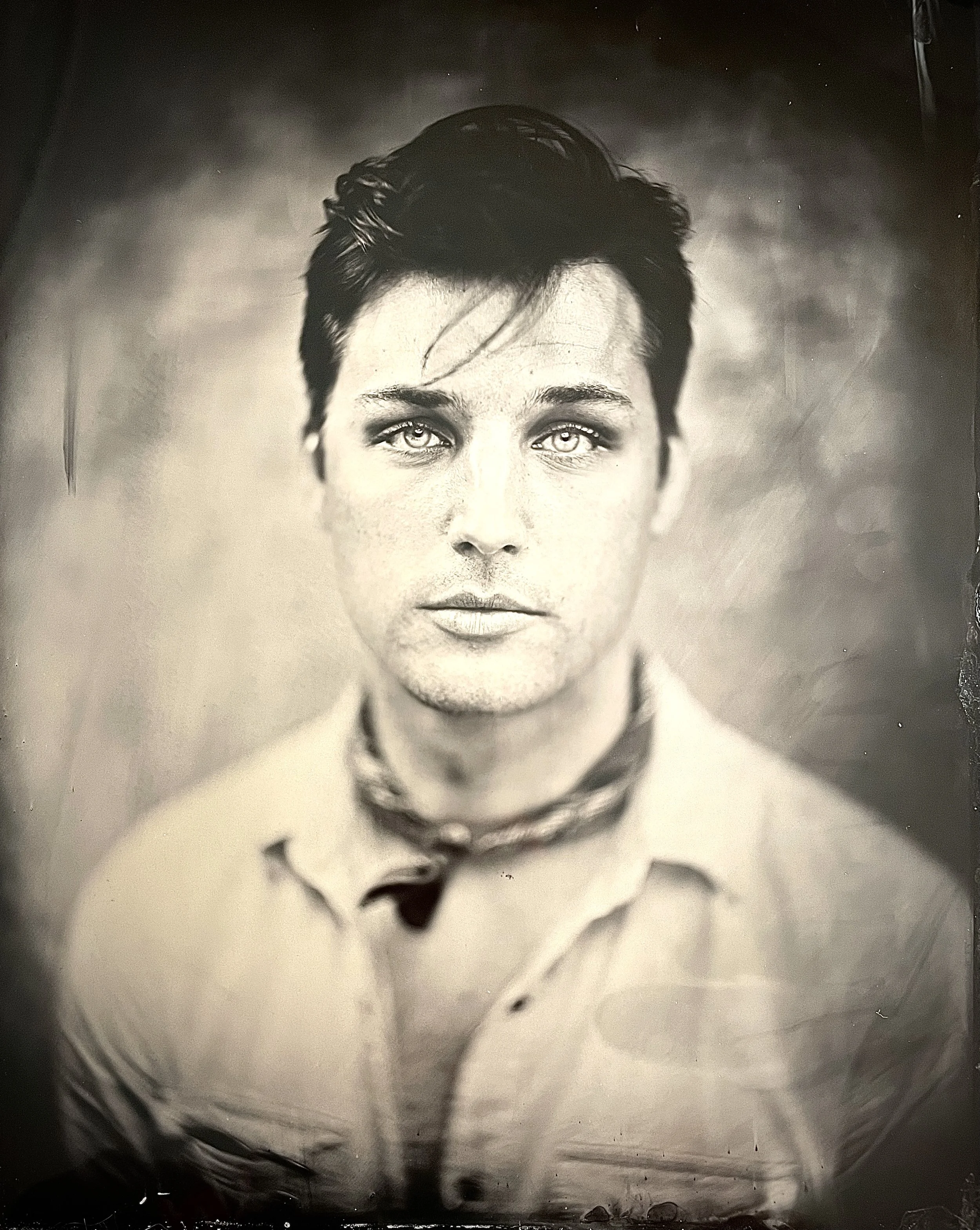 A black-and-white photo of a young man with short dark hair, light eyes, and a serious expression. He is wearing a collared shirt, and the photo is taken in a studio with a blurred background.