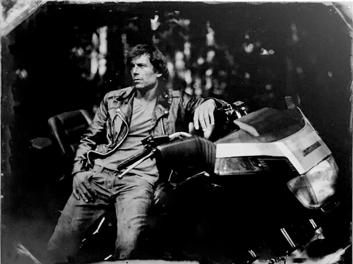 Black and white photograph of a man with curly hair, wearing a leather jacket and casual clothes, sitting on a motorcycle with a forest background.