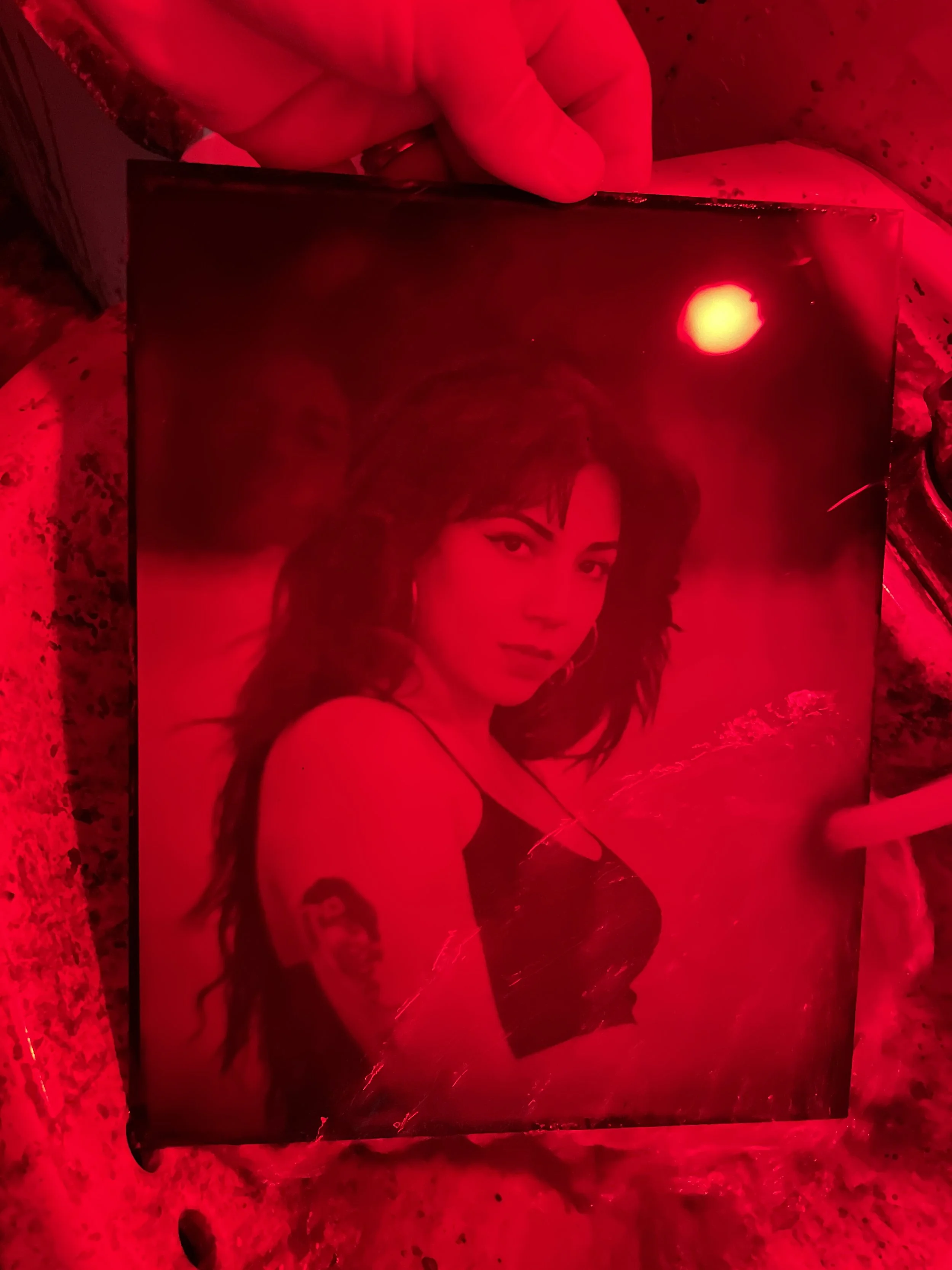 A photograph of a woman with dark, curly hair, wearing a black top, taking a selfie in a mirror illuminated by red light.