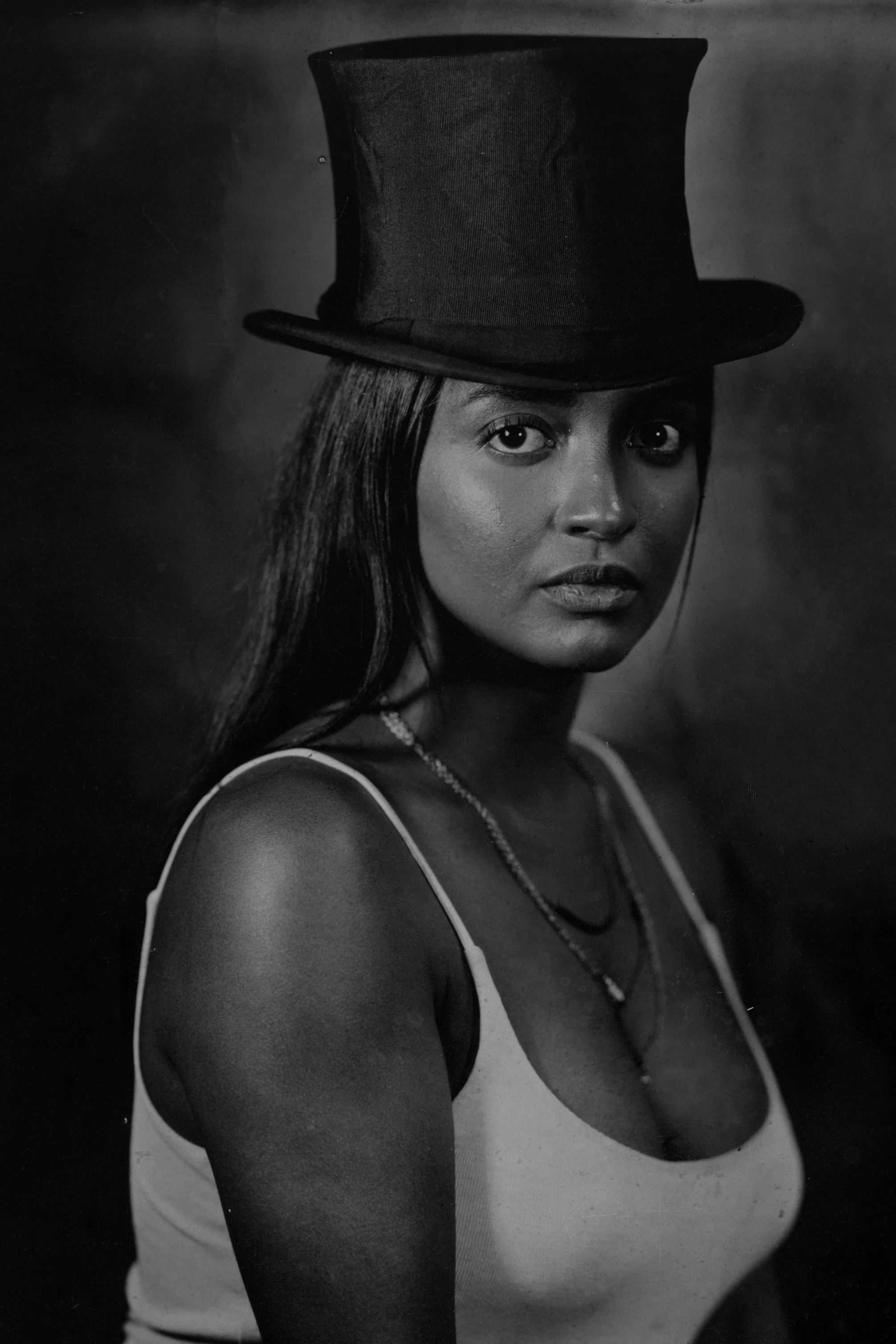 Black and white portrait of a woman wearing a top hat, with straight hair, wearing a sleeveless top, and looking directly at the camera.