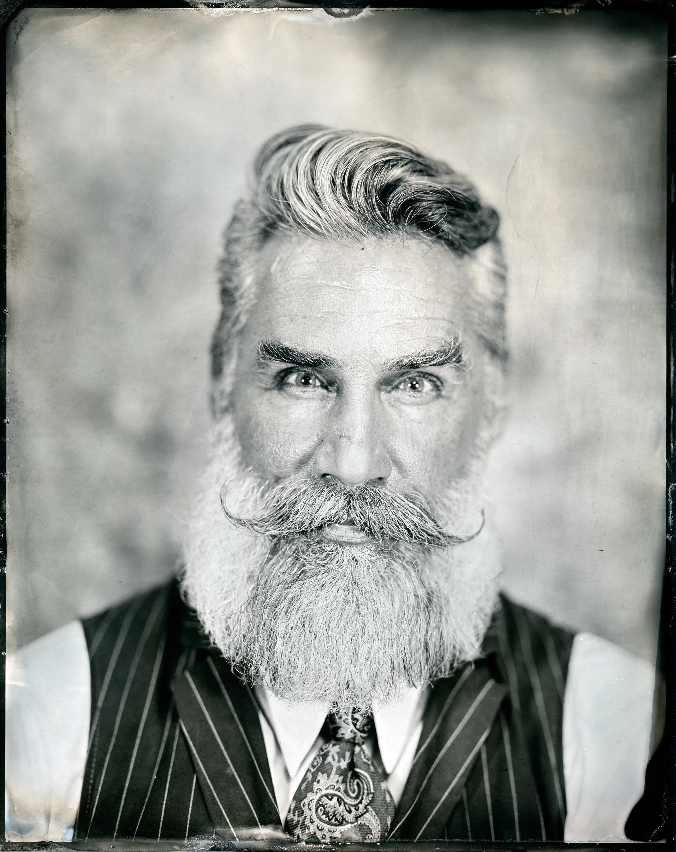 A black and white photograph of a man with a full beard and styled hair, wearing a pinstripe suit and a patterned tie, looking directly at the camera.