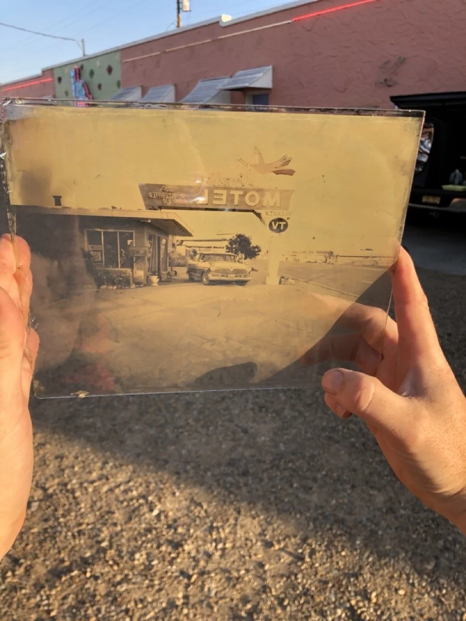Person holding a piece of dirty and cracked glass or plastic with a faded black-and-white photo of a vintage gas station, a car, and a sign that reads 'MOTEL' in front of a building; the background shows a brick building and a parking lot.