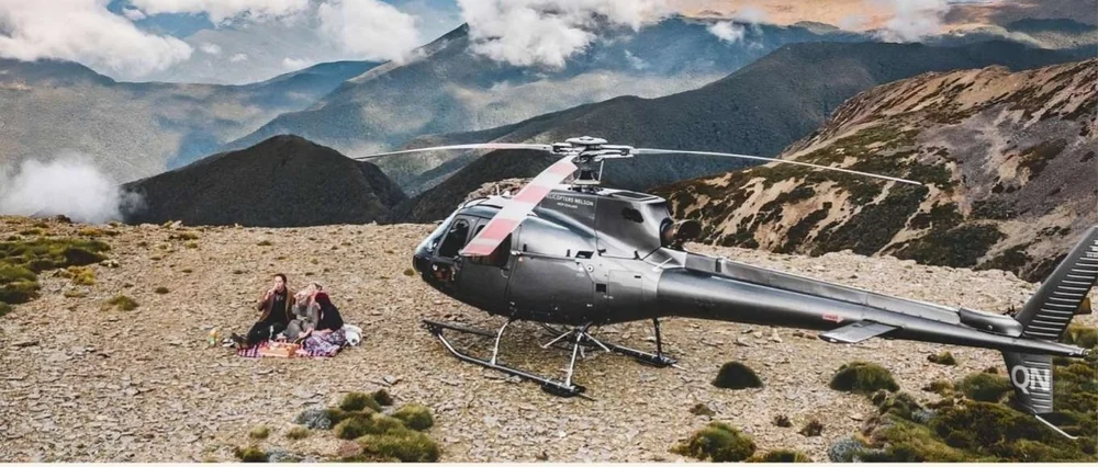 A grey helicopter landed on a mountaintop with guests picnicking beside it