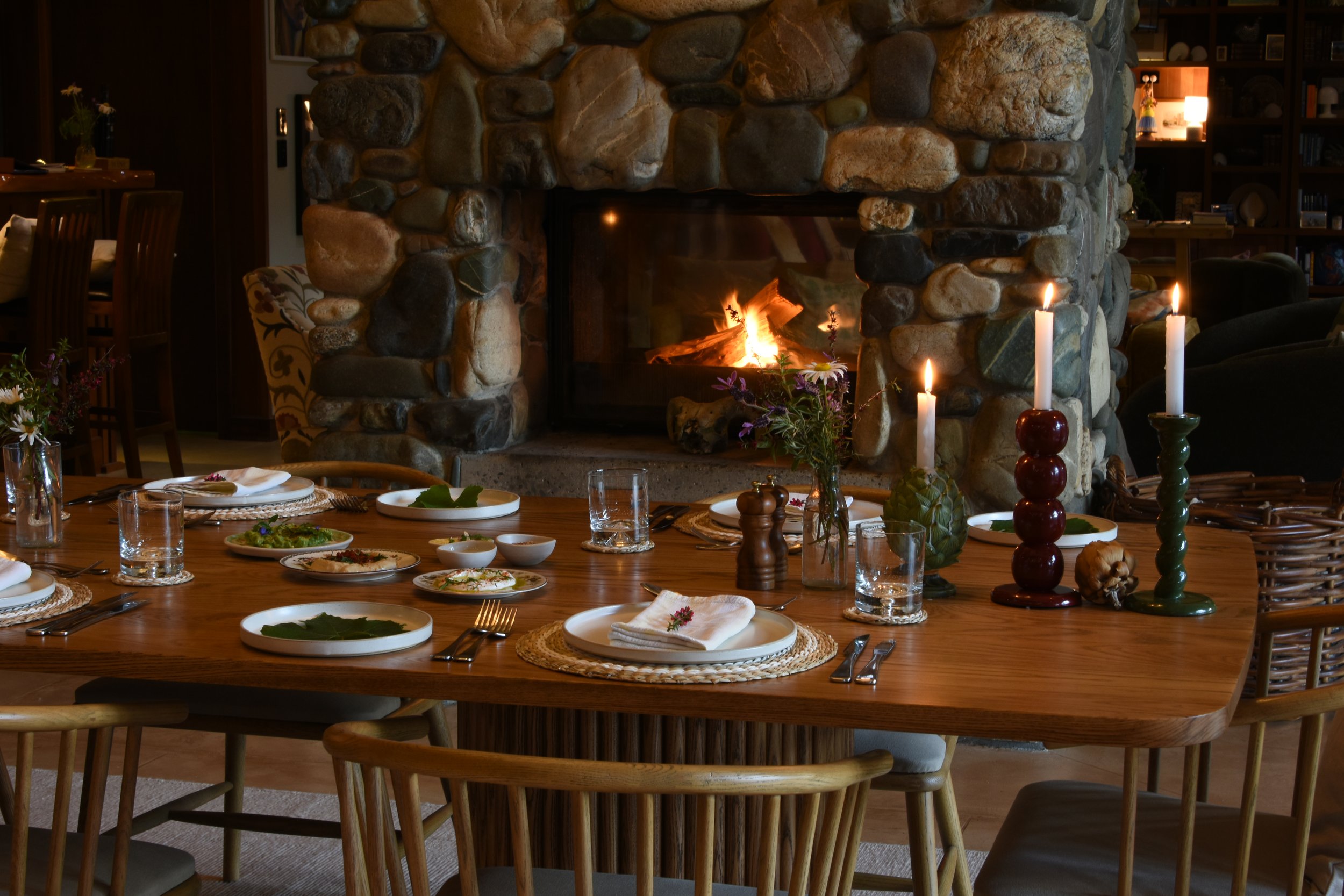 A cozy dining room with a large wooden table set with plates, glassware, and napkins, in front of a rustic stone fireplace with a burning fire. There are candles, flowers, and decorative table items, creating a warm atmosphere at Stonefly Lodge