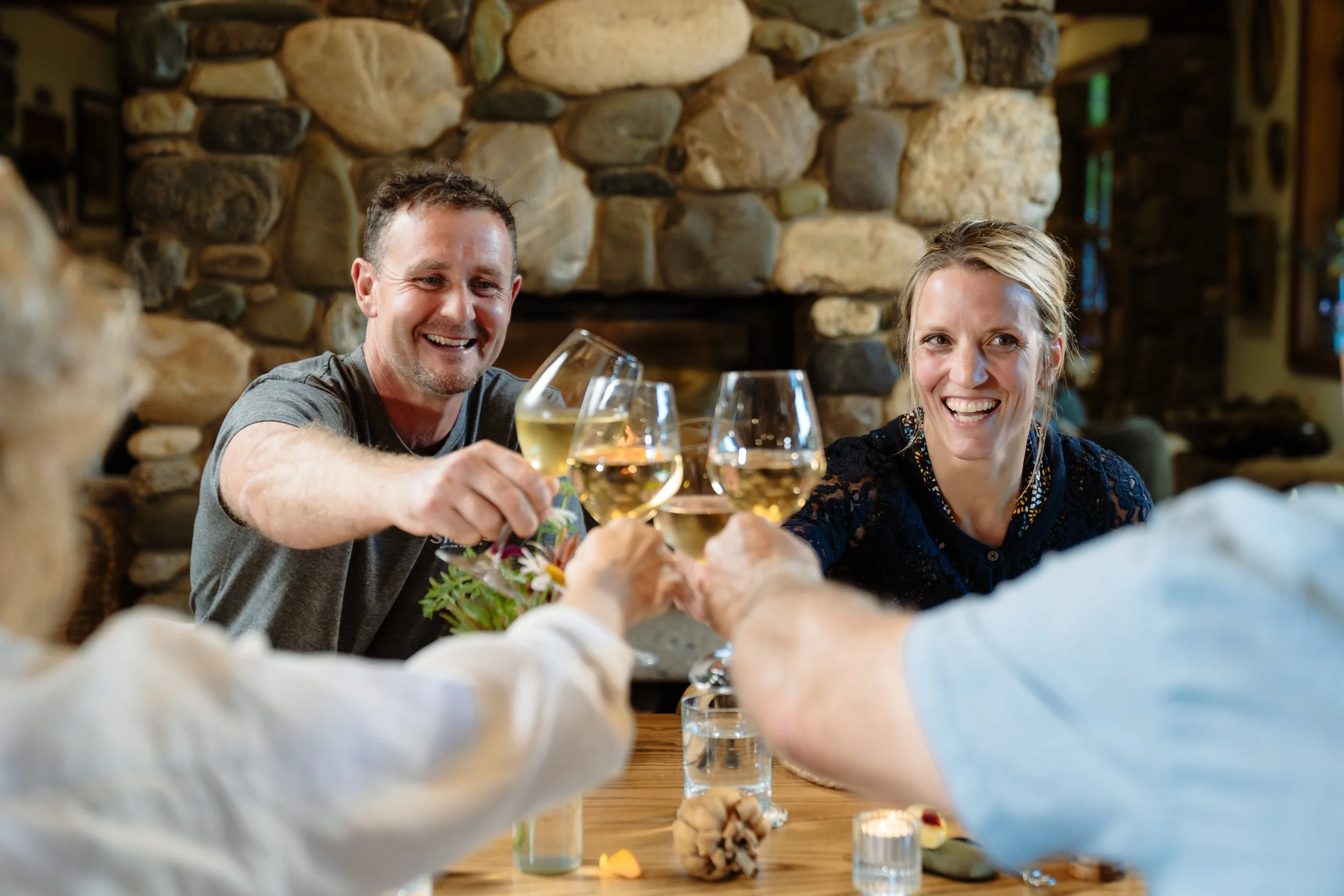 A group of four adults enjoying a toast with glasses of white wine at a rustic wooden table in a cozy room with a stone fireplace.