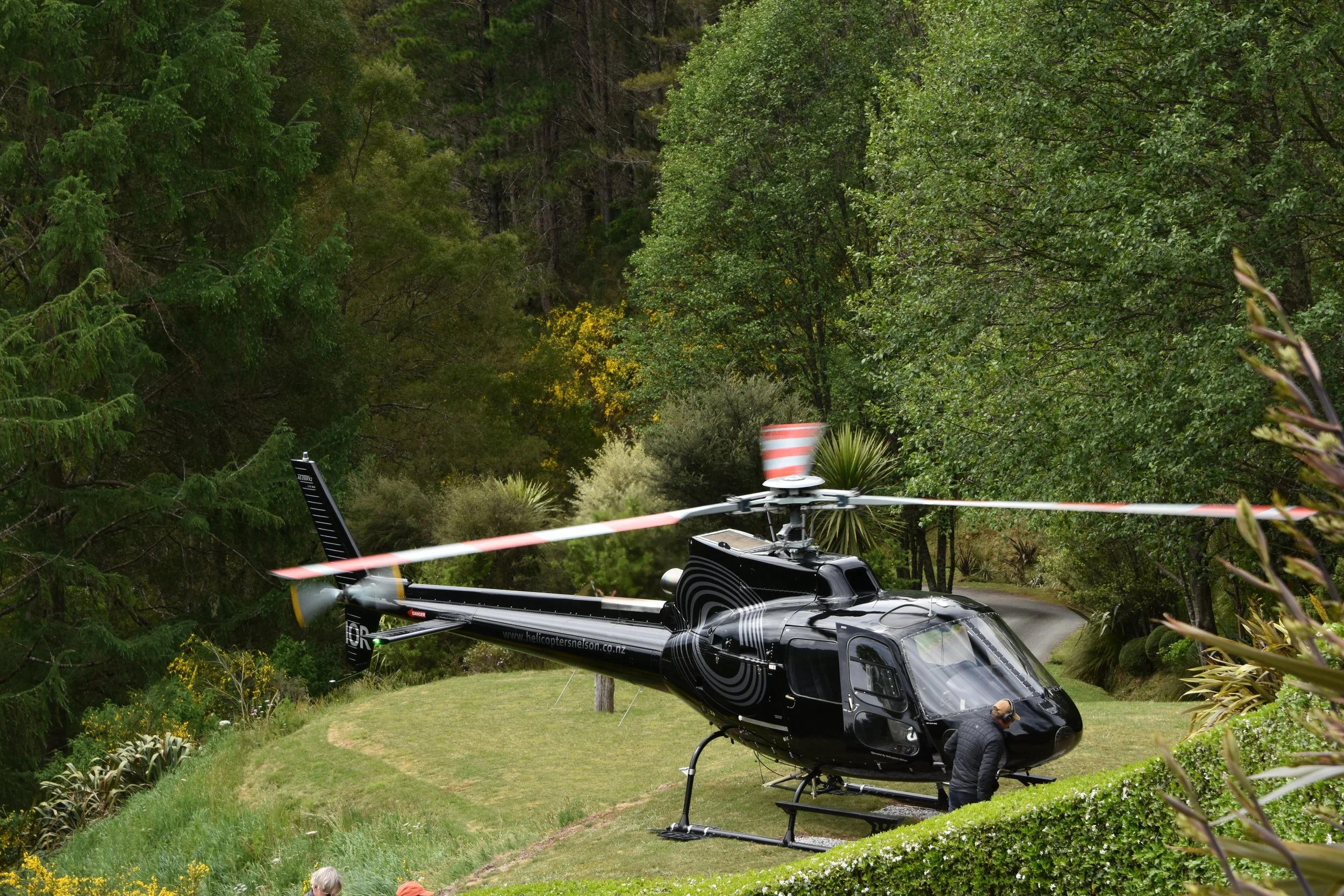 A black helicopter parked on the grassy Stonefly Lodge helipad surrounded by trees, with a person standing near the front of the helicopter and the rotor blades spinning.
