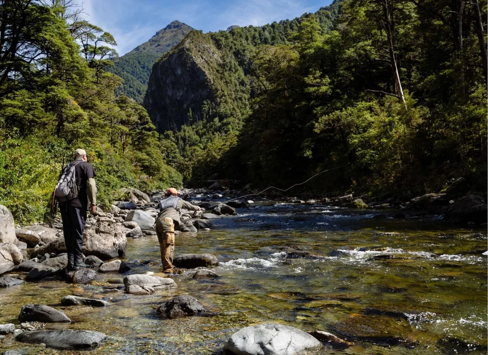 Two anglers working a clear boulder-strewn river below forested peaks