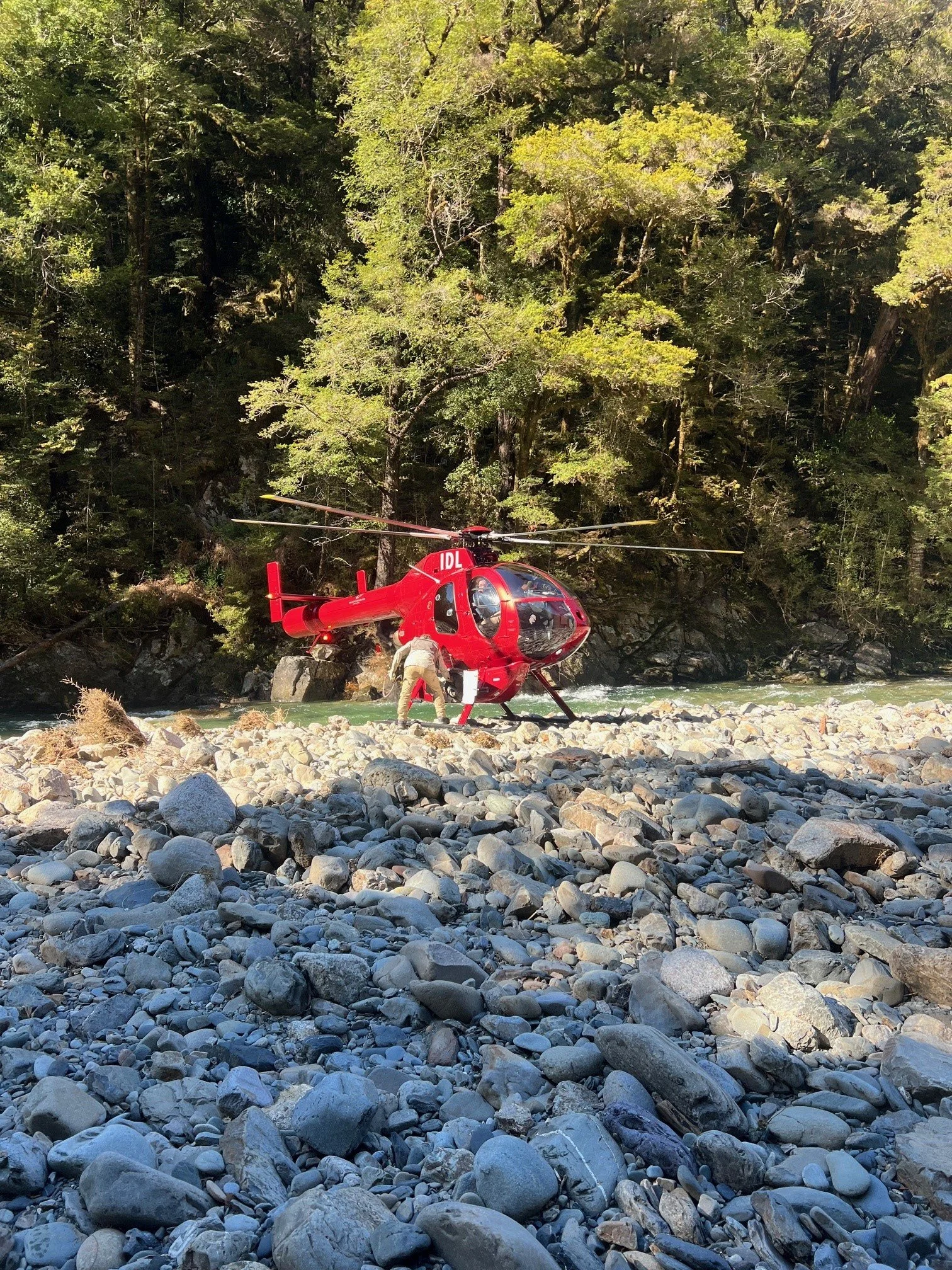 A red helicopter on rocky riverbank with trees in the background.