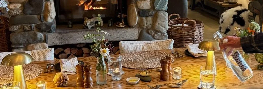 A cozy dining table setup in front of a stone fireplace with logs, featuring a vase with flowers, pepper and salt shakers, glasses of water, plates, a spoon, and a candle holder. A person is pouring a beverage into a glass on the right.