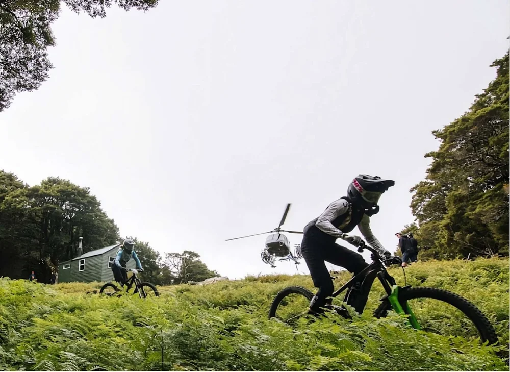A mountain biker riding through bracken with a helicopter and hut behind