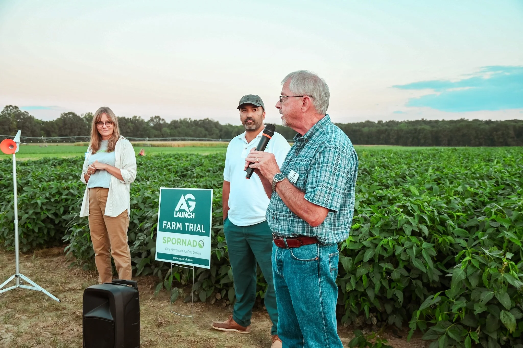 Three people standing outdoors in a farm field. The man in the front is speaking into a microphone, wearing glasses and a plaid shirt. The man in the middle is wearing a cap and a white shirt, looking towards the camera. The woman on the left is wear