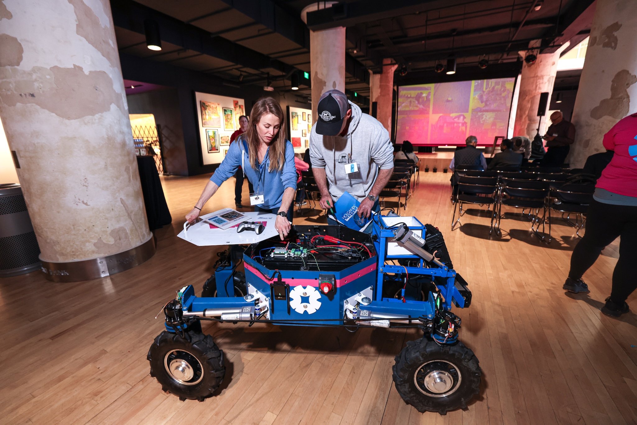 Two people working on a robot with wheels in an art gallery or exhibition space, with art displayed on the walls and a large screen in the background.