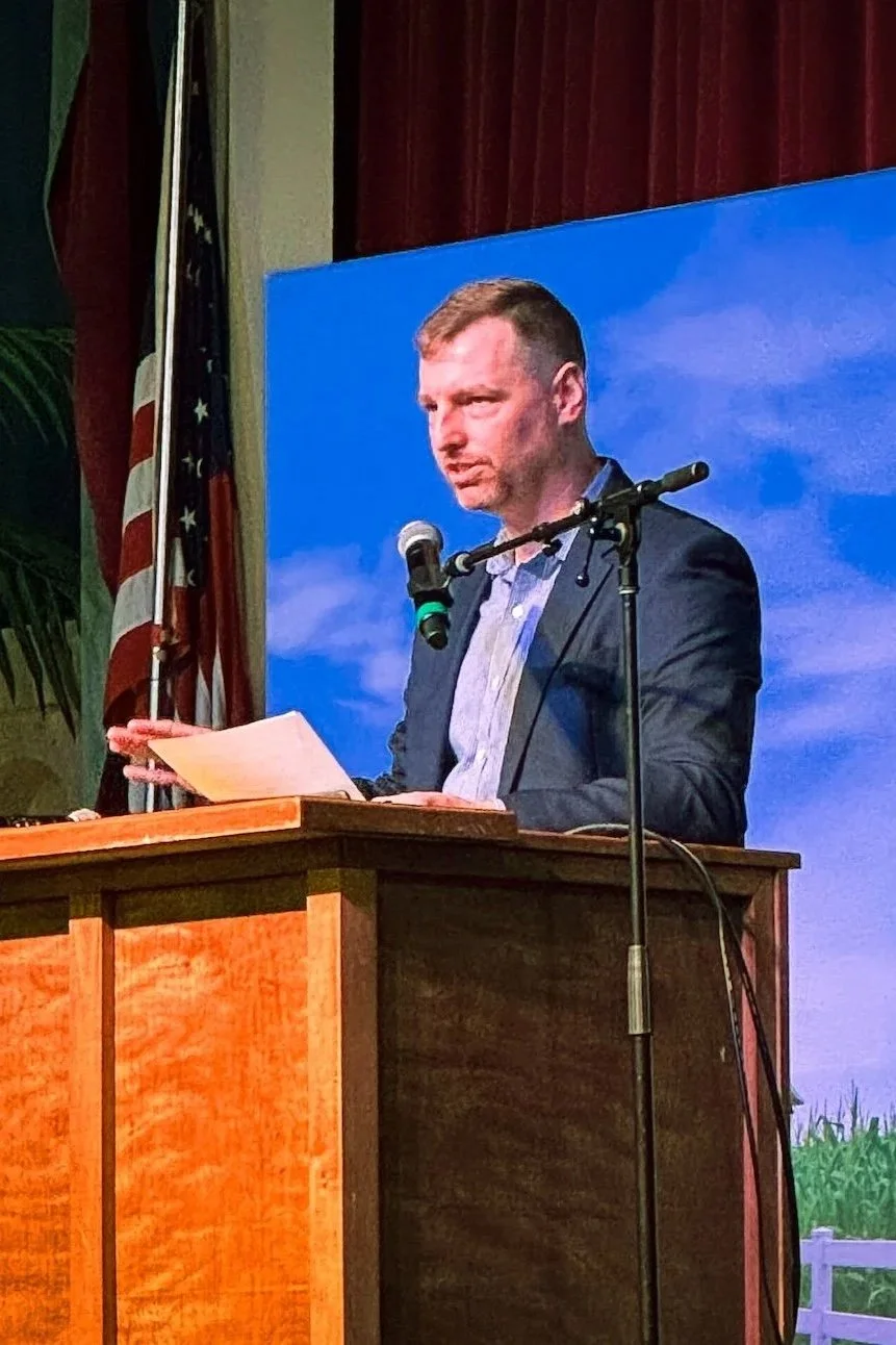 A man in a suit standing at a wooden podium with papers in hand, speaking into a microphone, with an American flag and a blue sky backdrop behind him.