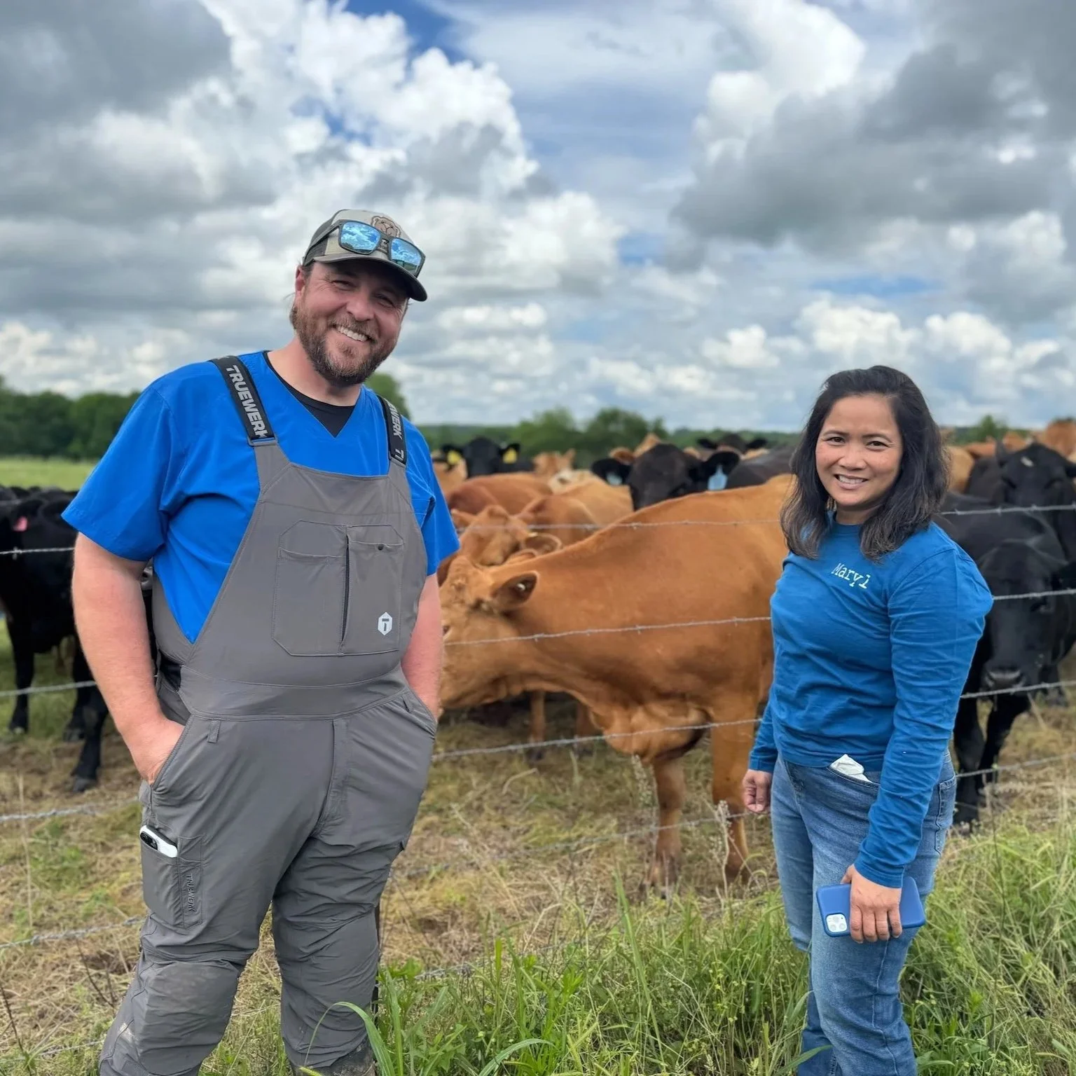 A man and woman standing outdoors in front of a cow pasture with a cloudy sky. The man is wearing gray overalls and a blue shirt, and the woman is wearing a blue shirt with the name 'Mary' on it and jeans. The man is smiling with his hands in his poc