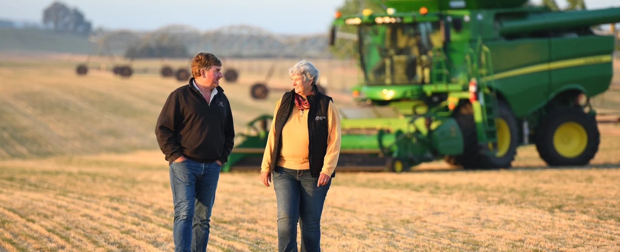 A man and a woman walking and talking in a field with farm machinery and a tractor in the background during daytime.