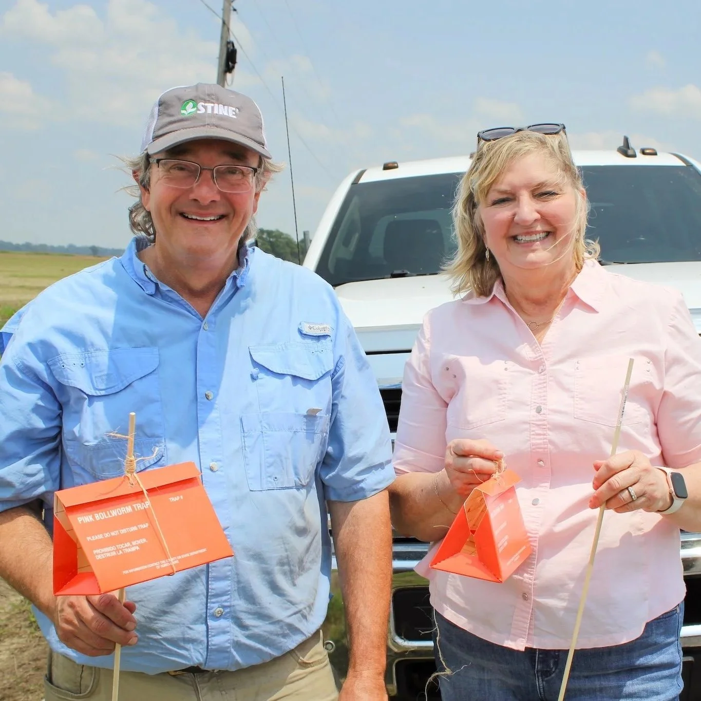 A smiling man wearing glasses, a gray cap, and a light blue shirt, standing next to a smiling woman wearing a light pink shirt and sunglasses on her head, both holding orange paper bags attached to sticks, outdoors with a white vehicle and open field