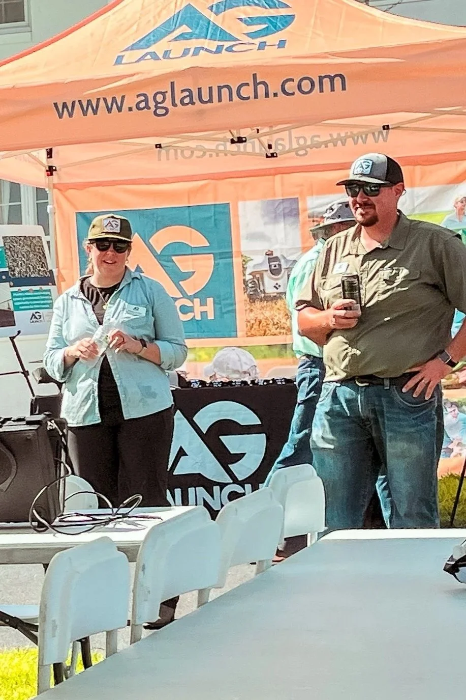 Two people standing outdoors in front of an orange tent with AG LAUNCH branding, at what appears to be an event or promotion. One person is wearing a light blue jacket and cap, holding a clear cup, and the other is in a green shirt and baseball cap, 