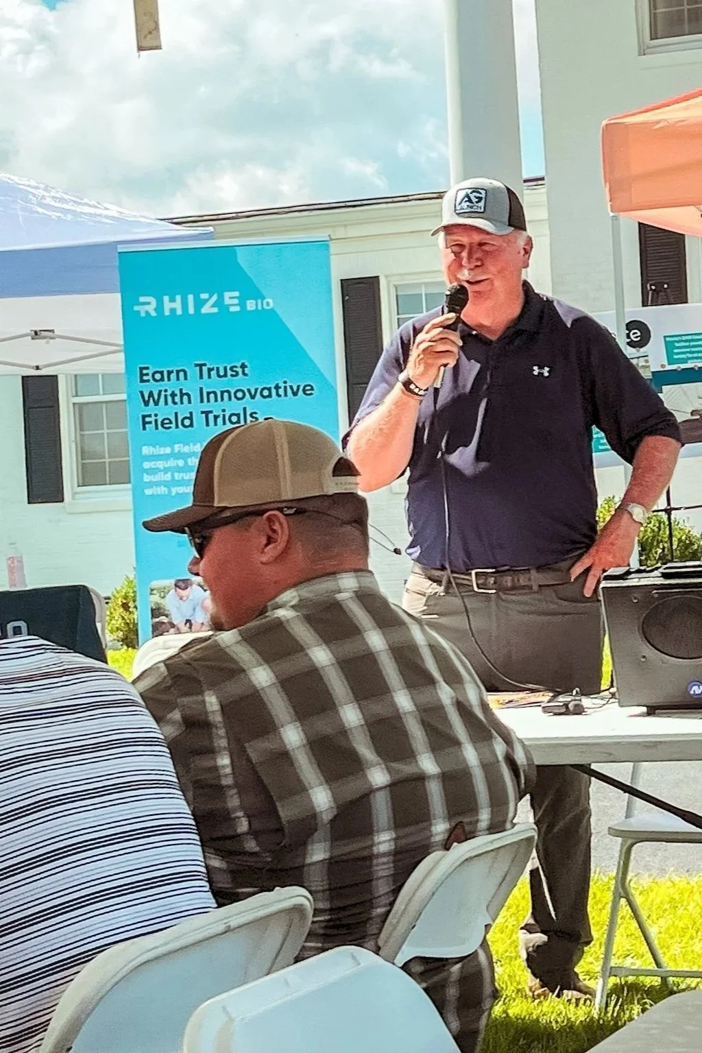 A man is speaking into a microphone at an outdoor event, with a banner that reads 'Rhize Bio' and 'Earn Trust With Innovative Field Trials' in the background. Several other people are seated at tables under a tent with a house visible behind.