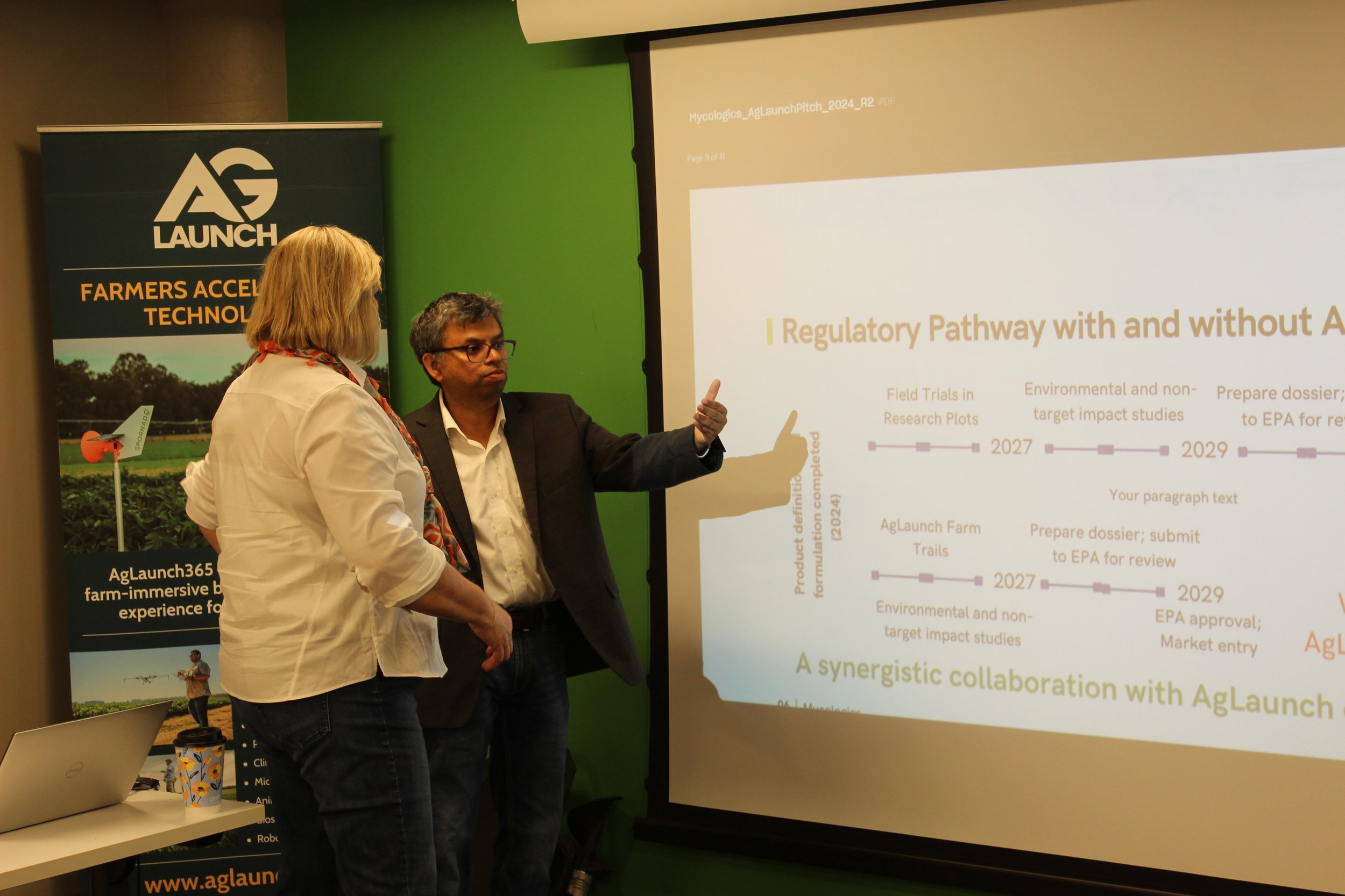 Two individuals are standing in front of a presentation screen. The woman in the foreground has blonde hair and wears a white blazer, while the man is wearing a dark blazer and glasses. They appear to be engaged in discussion during a conference or s