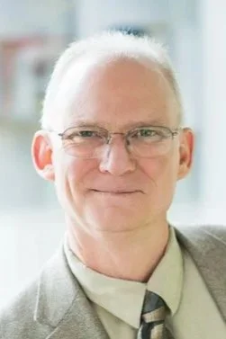 Portrait of an elderly man with white hair, glasses, and a light-colored suit, outdoors with a blurred background.