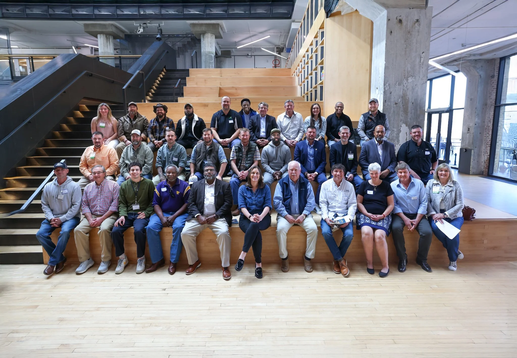 Group of diverse people sitting and standing on a wooden staircase inside a modern building with large windows and industrial design elements.