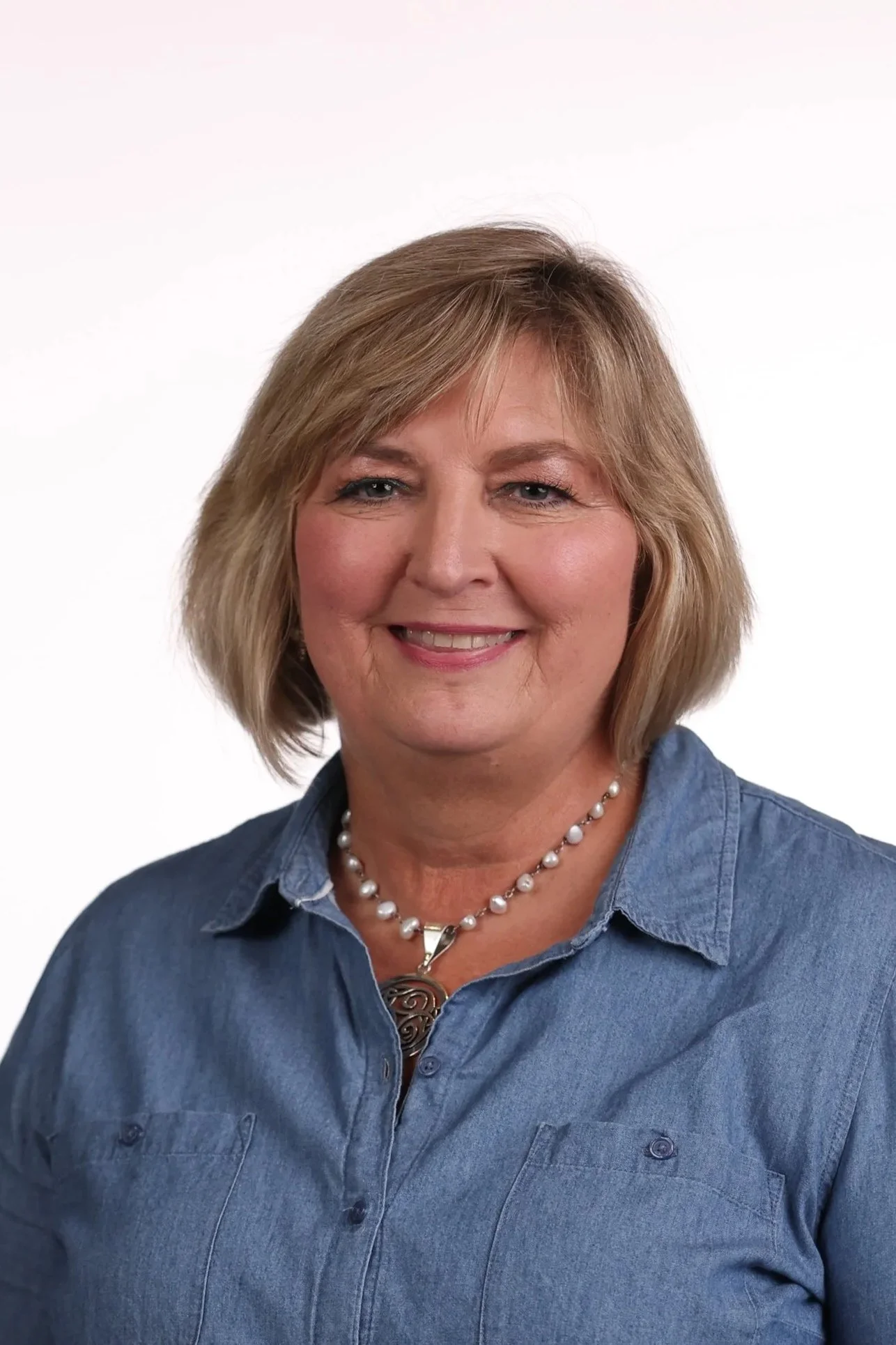 A middle-aged woman with shoulder-length blonde hair wearing a denim shirt and a pearl necklace, smiling at the camera against a plain white background.